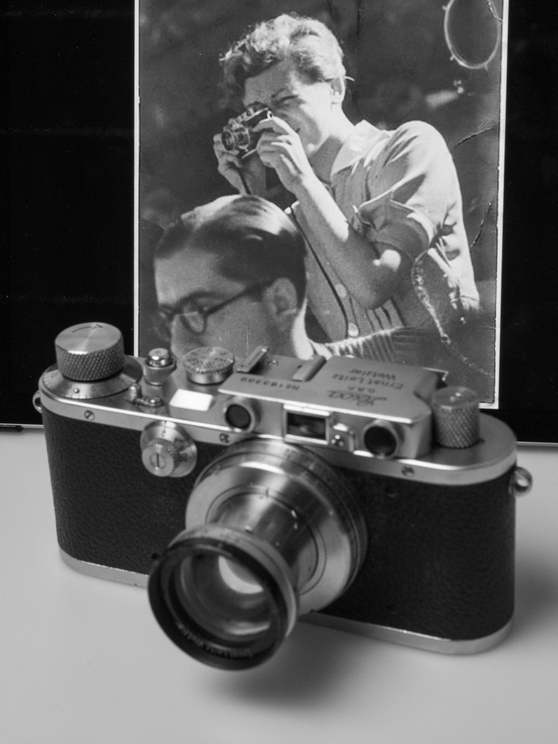Note the strap on the camera that Gerda Taro is using. Lugs for it were introduced only with the third iteration of the mass-produced screw-mount Leica, the Model III from 1933. The attached lens has been identified as a Summar 5cm; exactly the lens that is on the camera in front