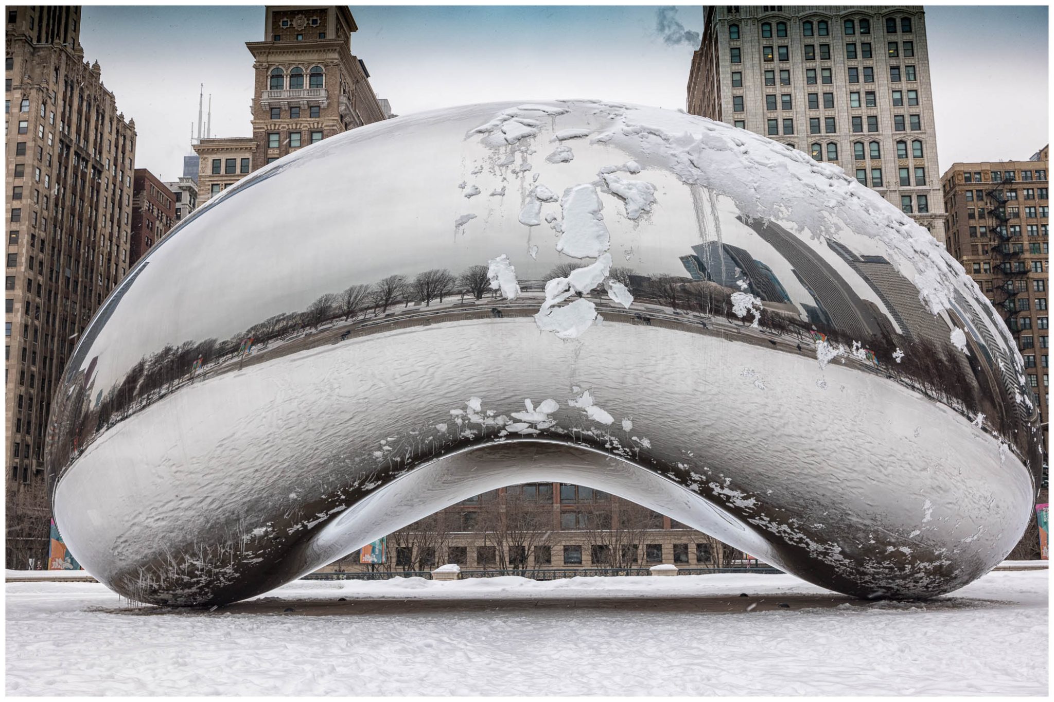 A close-up of the Cloud Gate sculpture, commonly known as 'The Bean,' partially covered in snow, reflecting surrounding buildings in a winter setting.