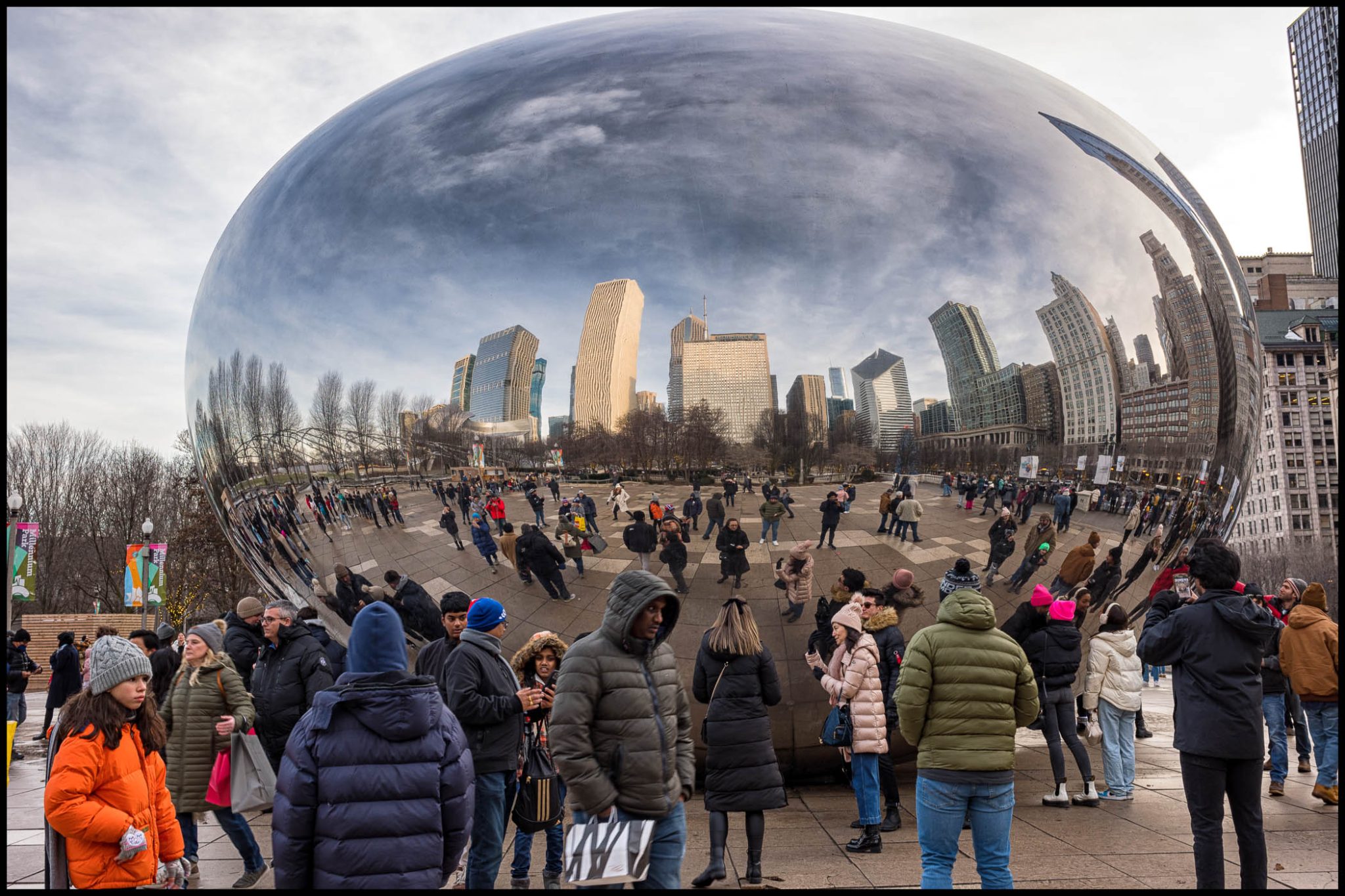 A large, reflective metallic sculpture, resembling a bean, surrounded by numerous people in an urban park with skyscrapers in the background.