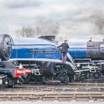 Two steam locomotives named 'Sir Nigel Gresley' and 'Tornado' are parked on a railway track. A train engineer in a black jacket is climbing on the 'Sir Nigel Gresley' locomotive, while another man in a coat is observing nearby. The sky is overcast.