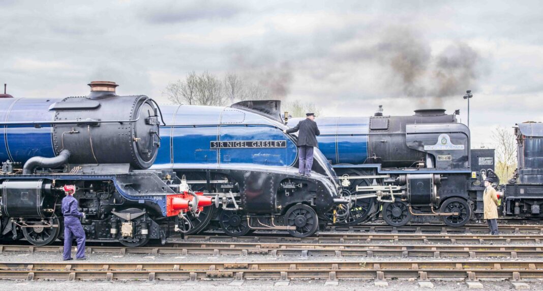 Two steam locomotives named 'Sir Nigel Gresley' and 'Tornado' are parked on a railway track. A train engineer in a black jacket is climbing on the 'Sir Nigel Gresley' locomotive, while another man in a coat is observing nearby. The sky is overcast.