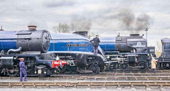 Two steam locomotives named 'Sir Nigel Gresley' and 'Tornado' are parked on a railway track. A train engineer in a black jacket is climbing on the 'Sir Nigel Gresley' locomotive, while another man in a coat is observing nearby. The sky is overcast.