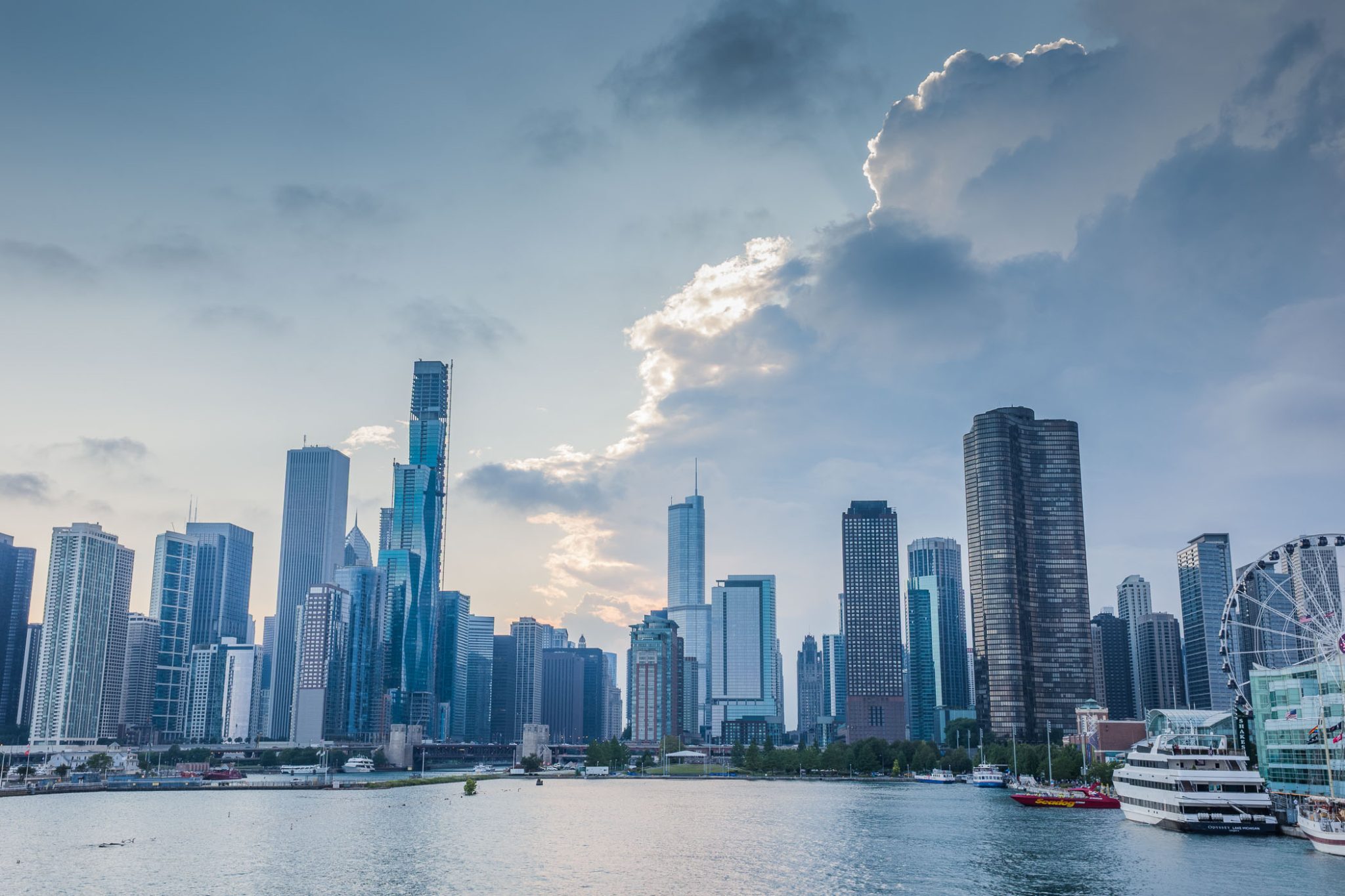 A panoramic view of a modern city skyline with tall skyscrapers, under a partly cloudy sky, alongside a body of water.
