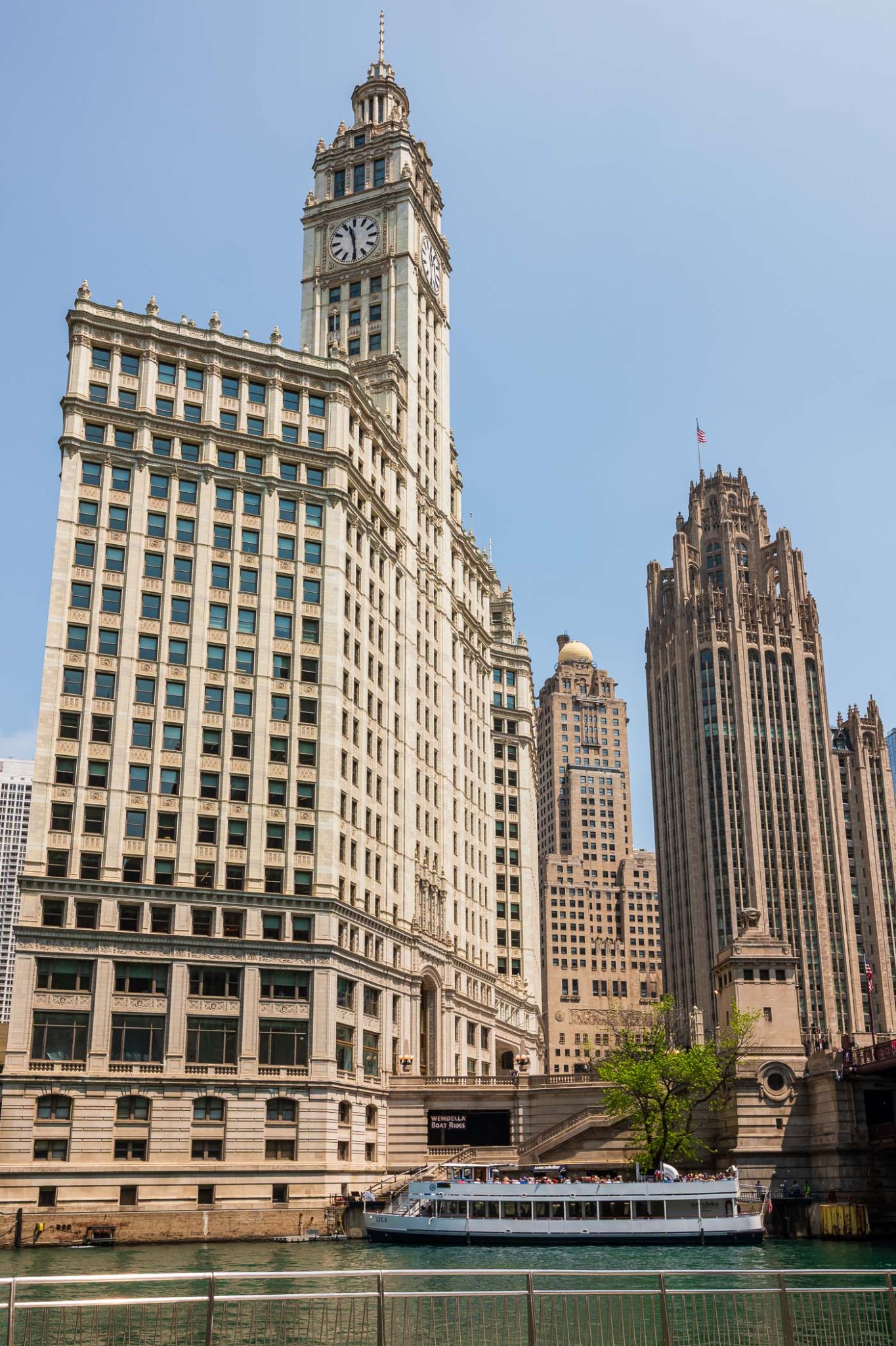 A view of the Chicago River featuring the historic Wrigley Building and Tribune Tower, with a sightseeing boat docked nearby.