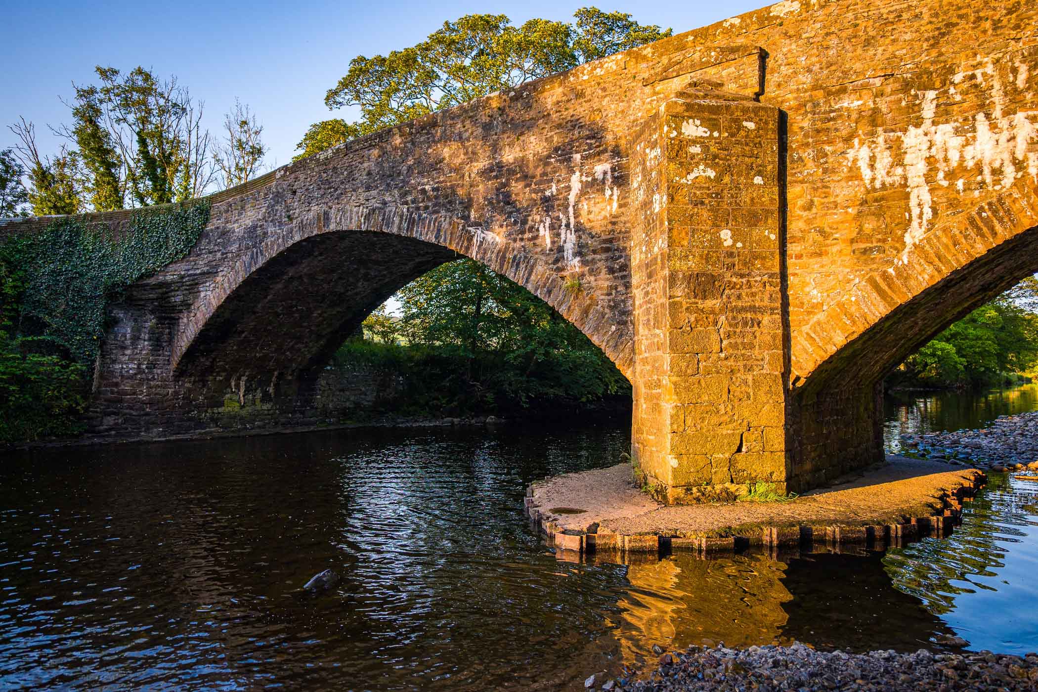 A stone bridge arching over a tranquil river, with lush greenery and reflections in the water, capturing the serene beauty of the English countryside.