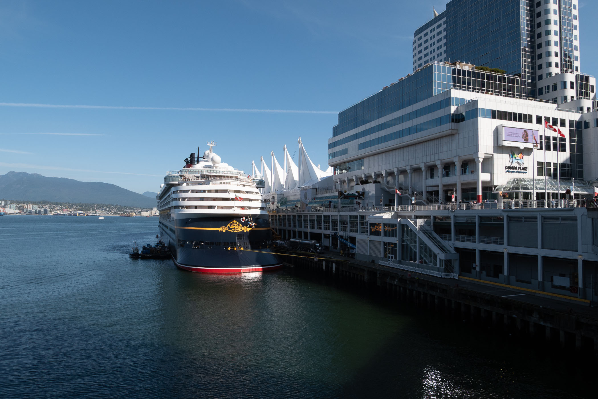 A large ship docked at the Vancouver harbor with a modern building and mountain backdrop under a clear blue sky.
