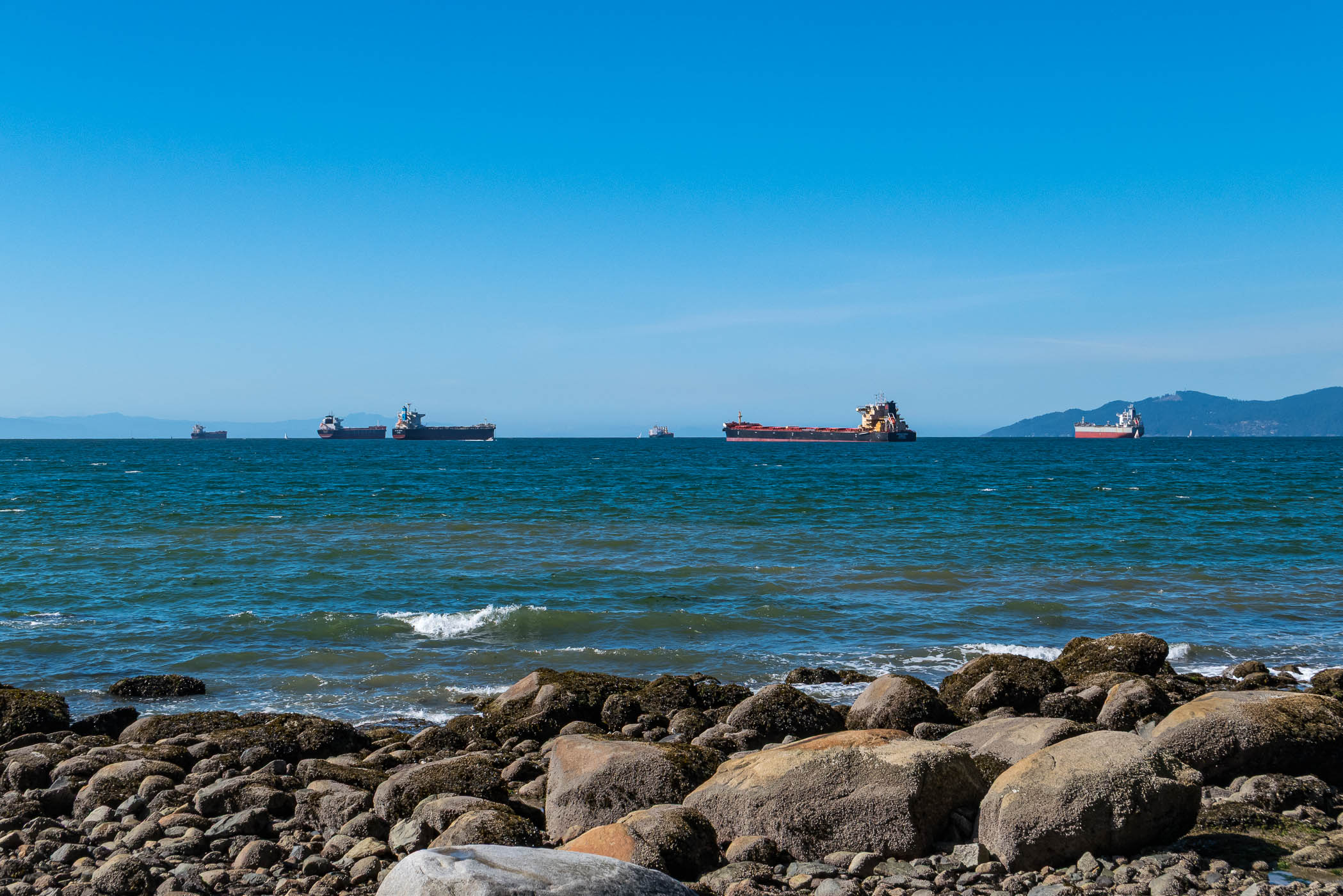 View of ships anchored in the ocean near rocky shoreline under a clear blue sky.