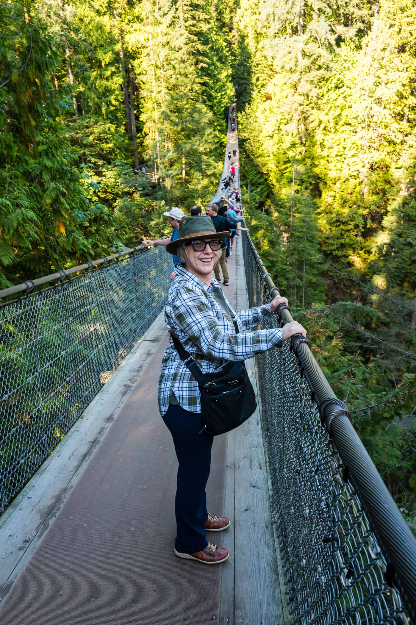 A woman stands on a suspension bridge surrounded by lush green trees, smiling while holding onto the railing.