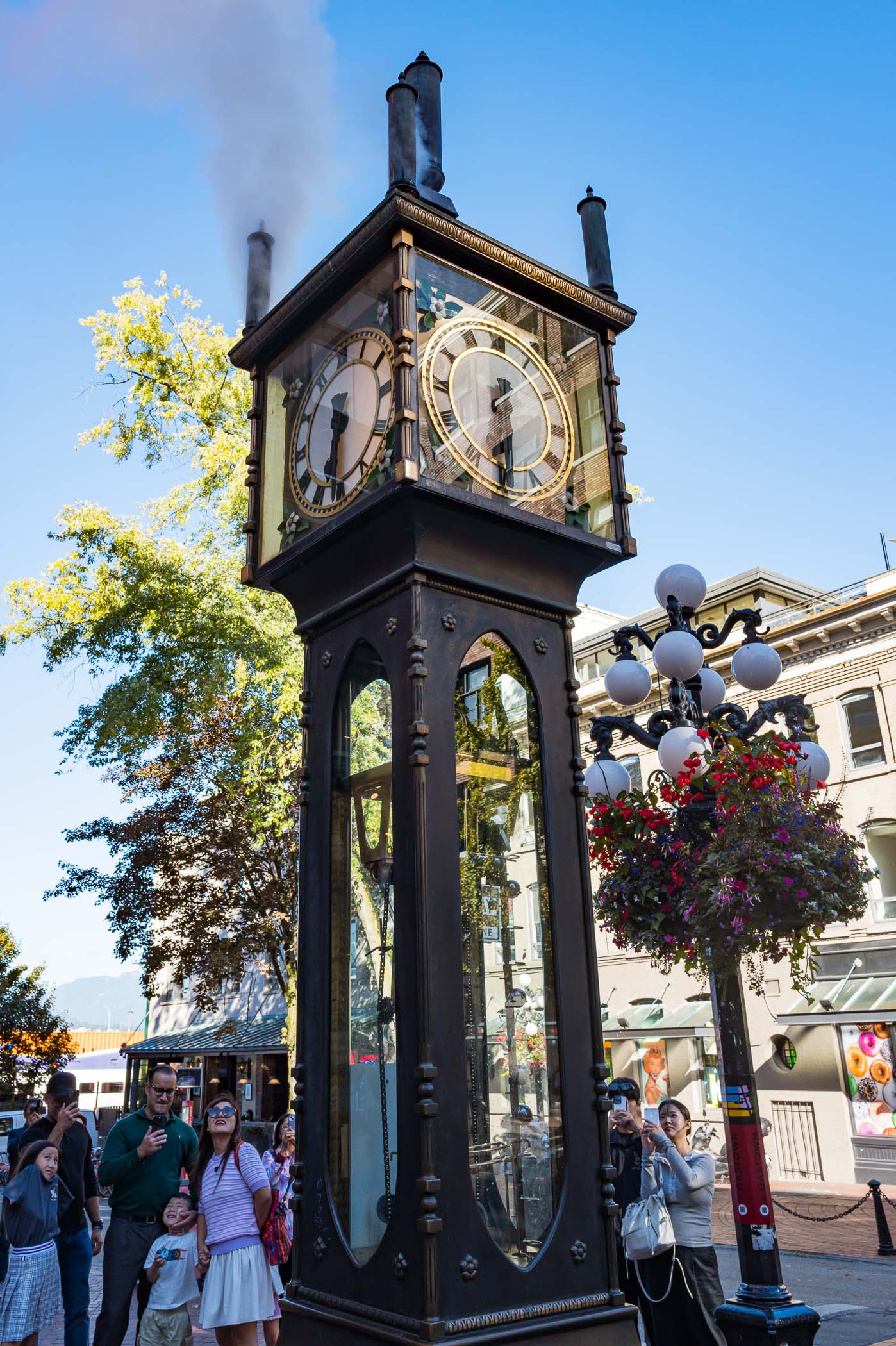 A steam clock in Gastown, Vancouver, displaying its time with smoke billowing from the top, surrounded by a group of people, including children.