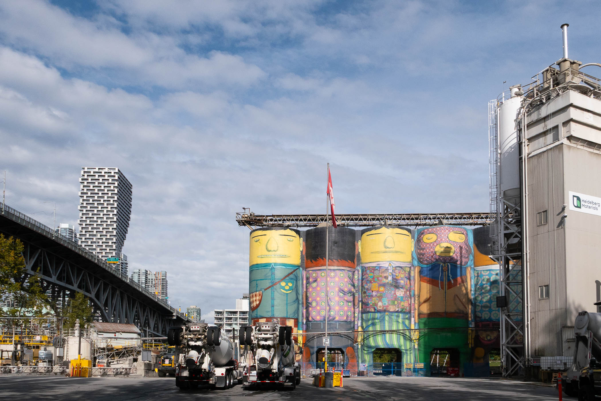 Colorful mural on silos in an industrial area, with concrete trucks in the foreground and a modern building in the background under a partly cloudy sky.
