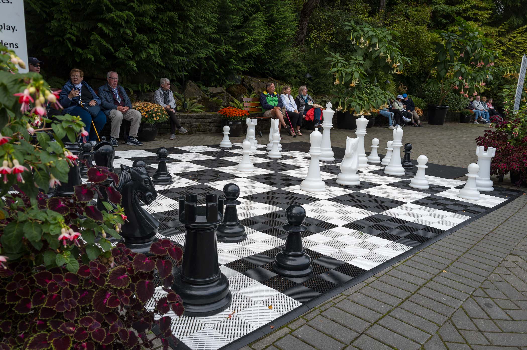 A large outdoor chess board with oversized black and white pieces, surrounded by colorful flowers and plants. Several people are seated in the background, watching the game in a garden setting.