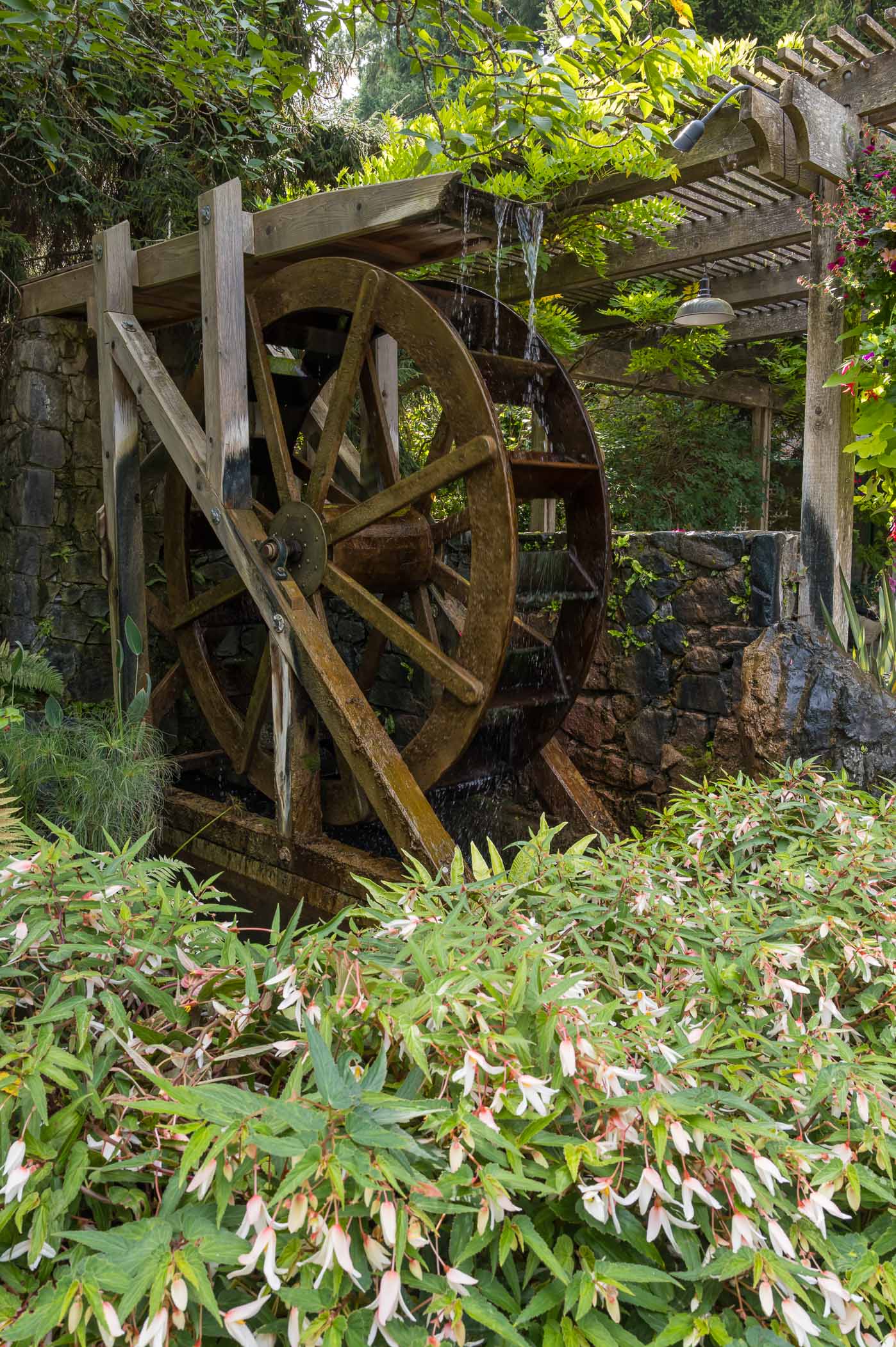 A wooden water wheel surrounded by lush greenery and flowering plants, with water cascading from the wheel into a stone structure.
