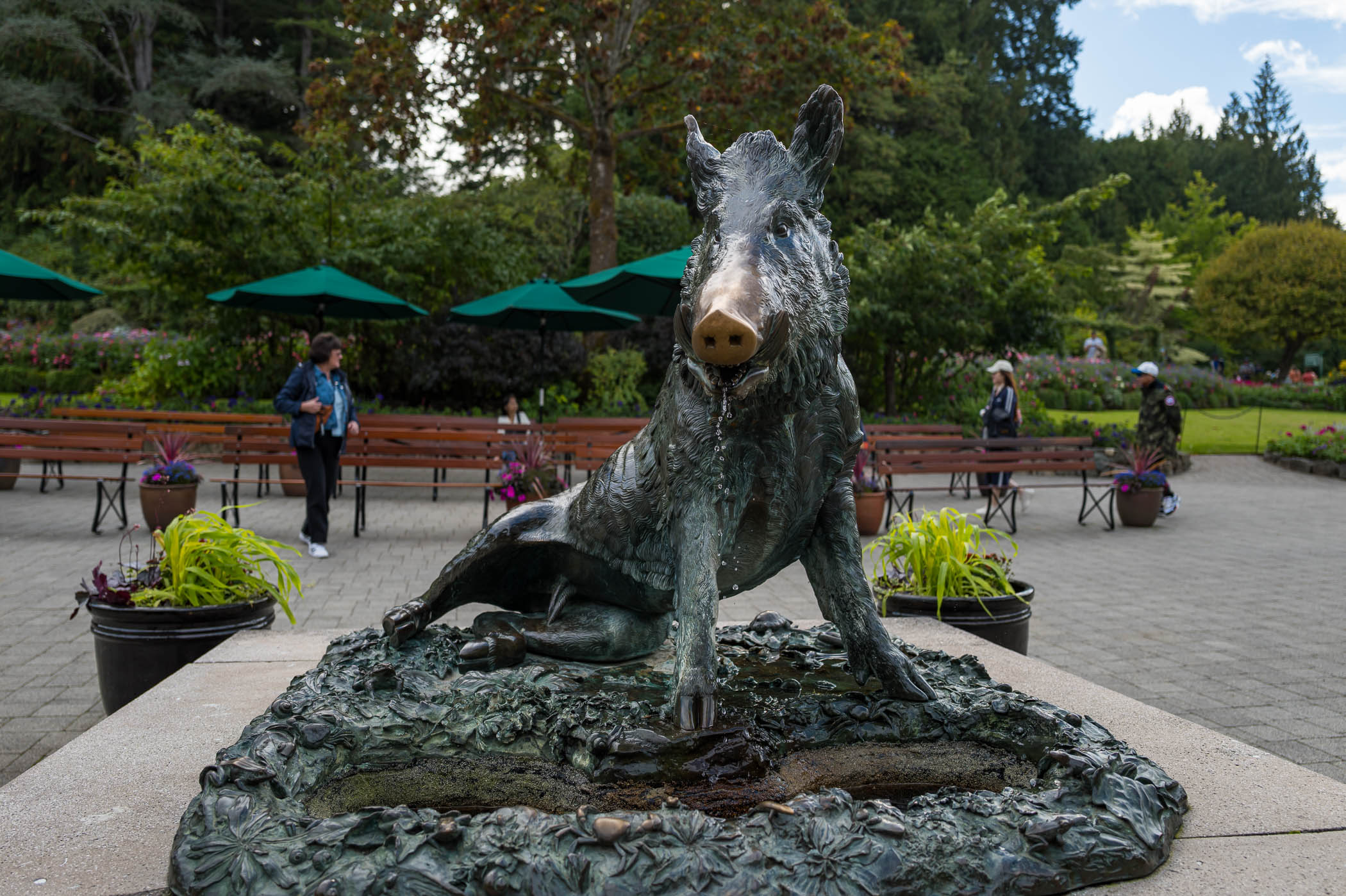 A bronze sculpture of a wild boar, situated in a garden area with greenery and benches in the background.