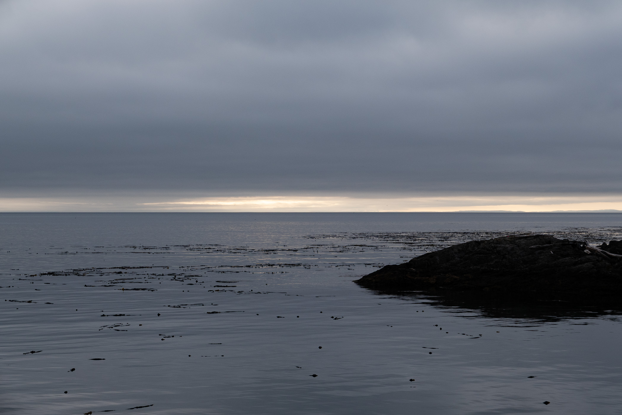 A cloudy sunset over a tranquil ocean, with gentle ripples in the water and a rocky shoreline in the foreground.