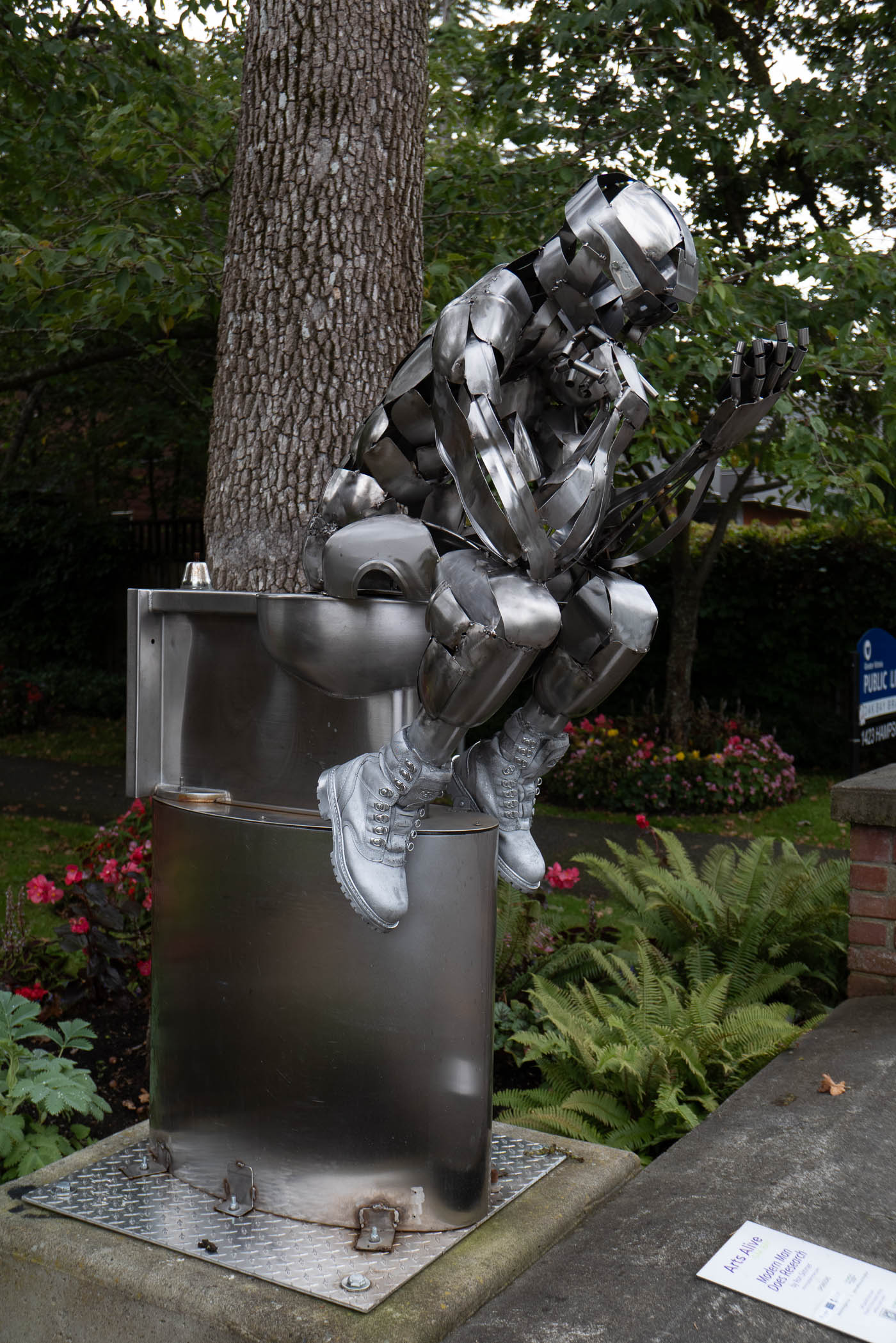 A metallic sculpture depicting a humanoid figure sitting on a shiny cylindrical base, with a tree in the background and colorful flowers surrounding it.