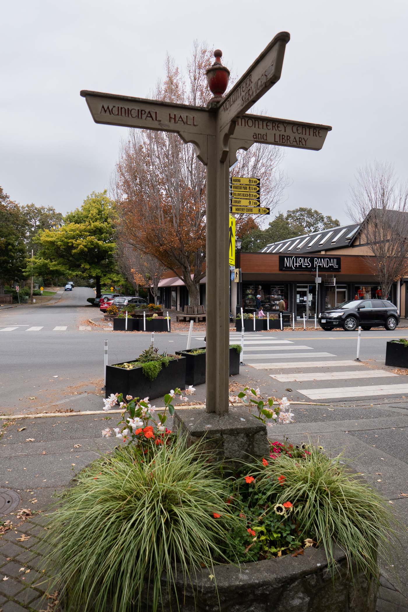 A directional signpost indicating various locations such as 'Municipal Hall' and 'Montery Centre and Library', surrounded by decorative plants and trees in an urban setting.