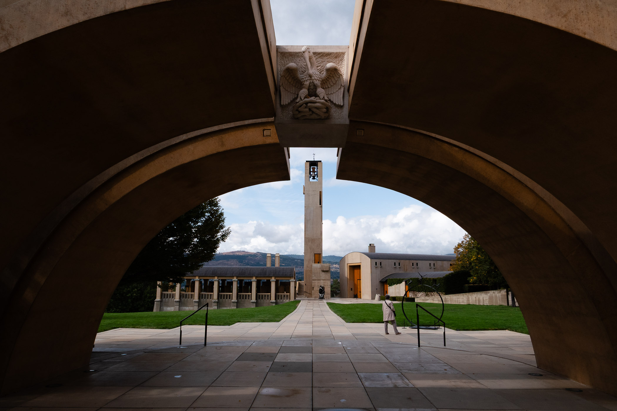 View of an architectural structure featuring arches, a bell tower, and a landscaped area, with a lone figure walking in the foreground.