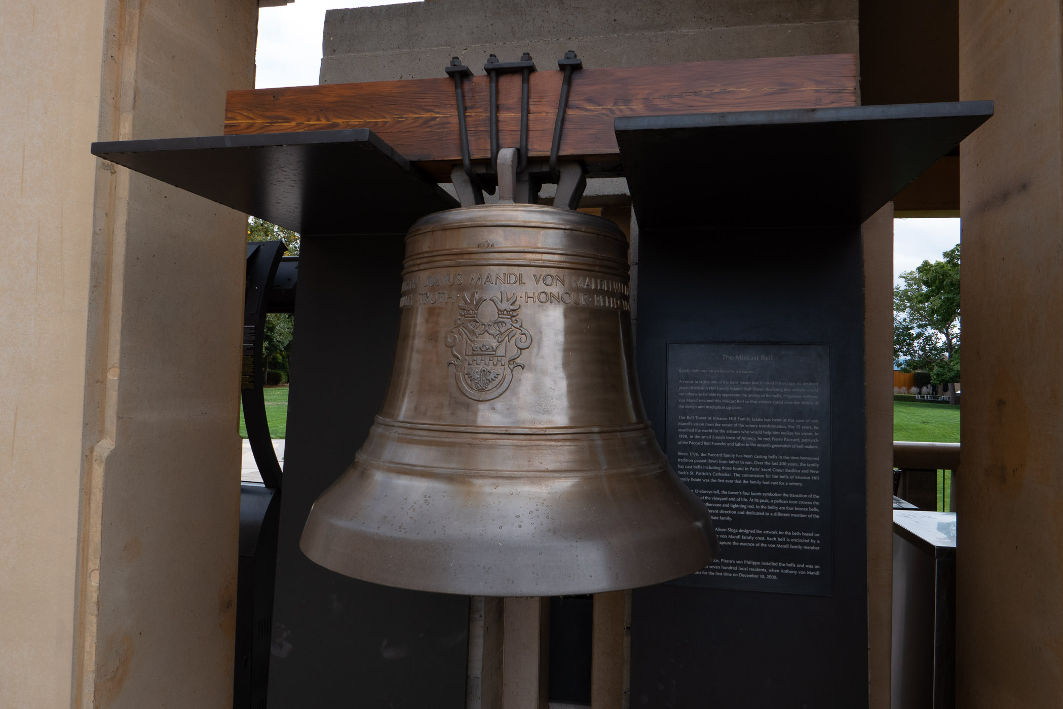 A close-up view of a large bronze bell mounted on a wooden and metal structure, with a plaque describing its history and significance in a public park.