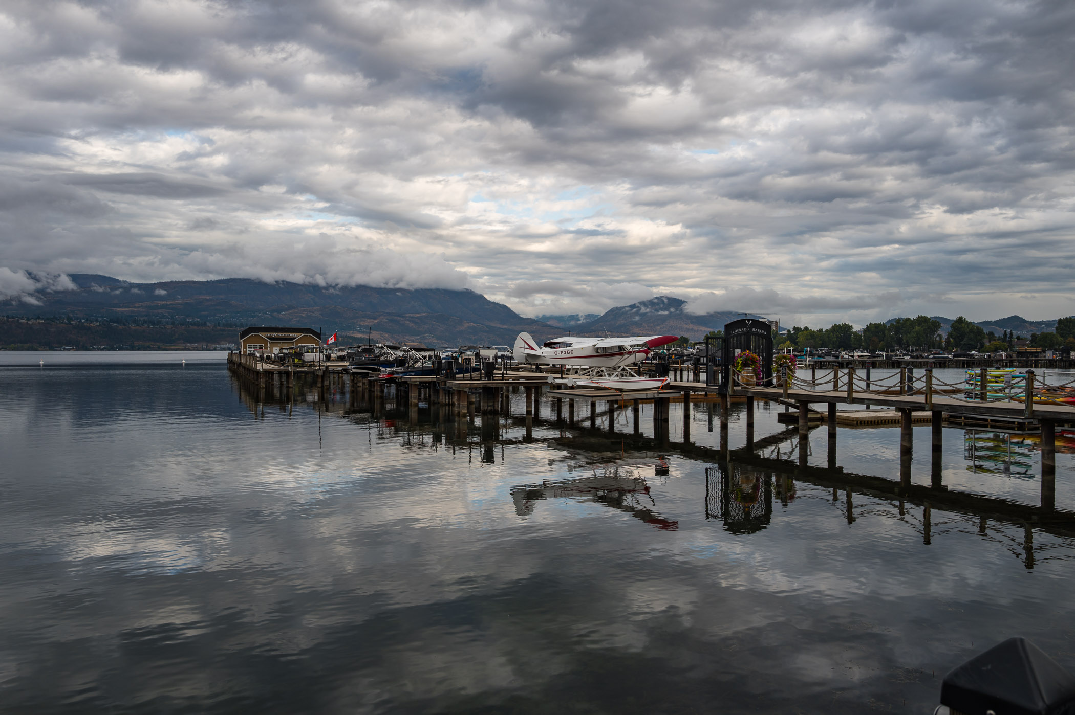 A peaceful lakeside view featuring a dock lined with boats and seaplanes, reflecting clouds and mountains in the calm water under a dramatic sky.