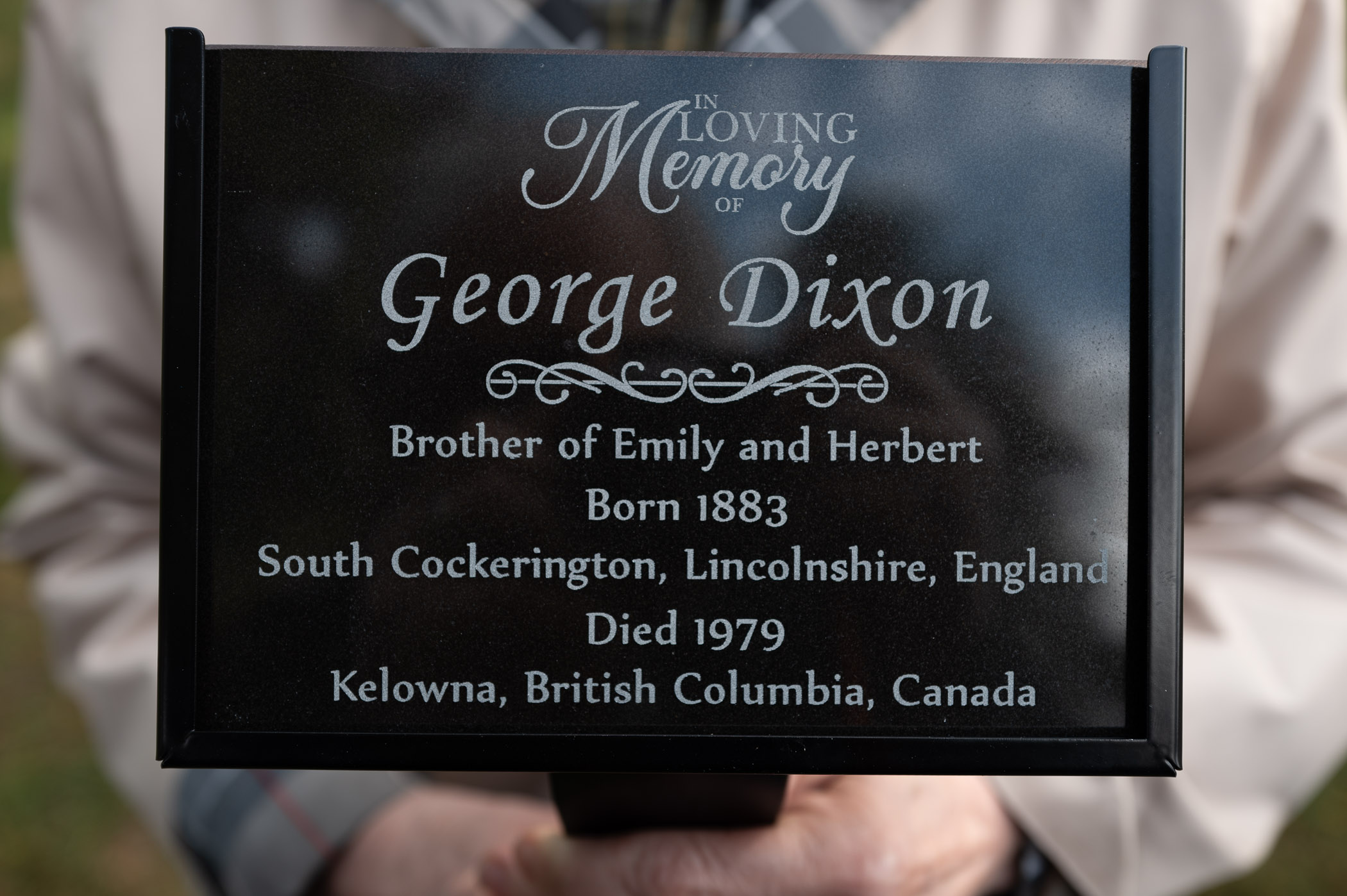 A close-up image of a gravestone plaque reading 'In Loving Memory of George Dixon. Brother of Emily and Herbert. Born 1883, South Cockerington, Lincolnshire, England. Died 1979, Kelowna, British Columbia, Canada.'