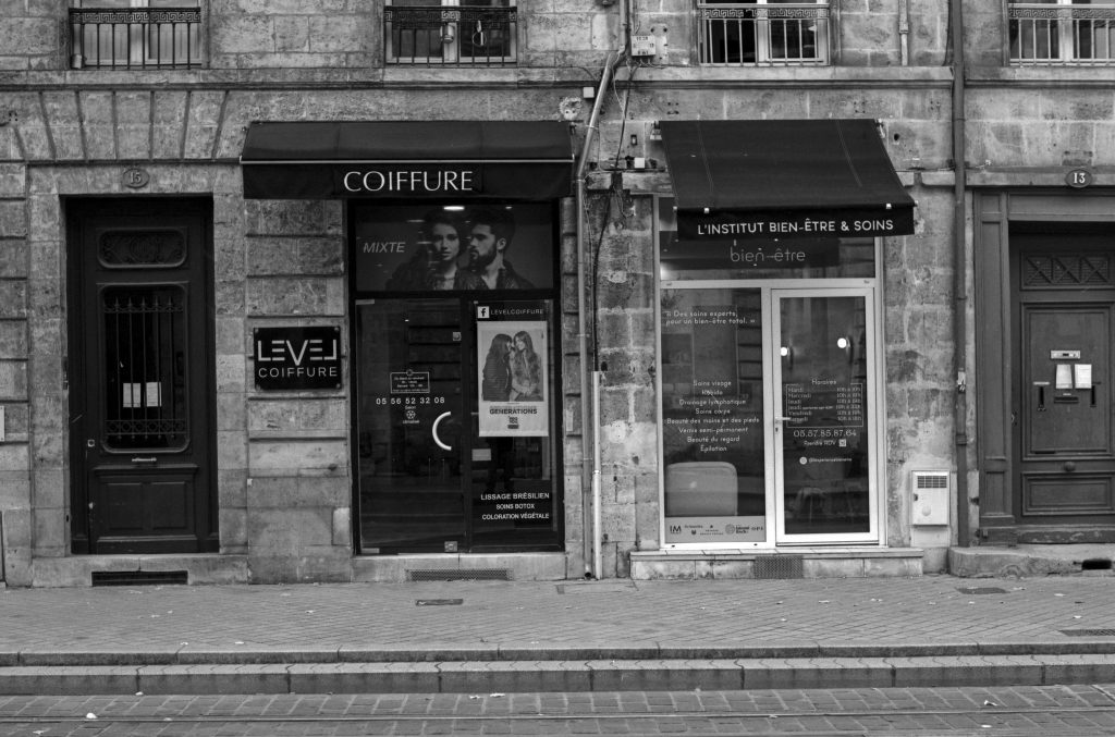 A black and white street view showcasing two adjacent shops with storefronts. On the left, 'LEVEL COIFFURE' has a modern design with a large window displaying hairstyles, and on the right, 'L'INSTITUT BIEN-ÊTRE & SOINS' features a glass front with service listings. Buildings with a stone facade and decorative balconies are visible in the background.
