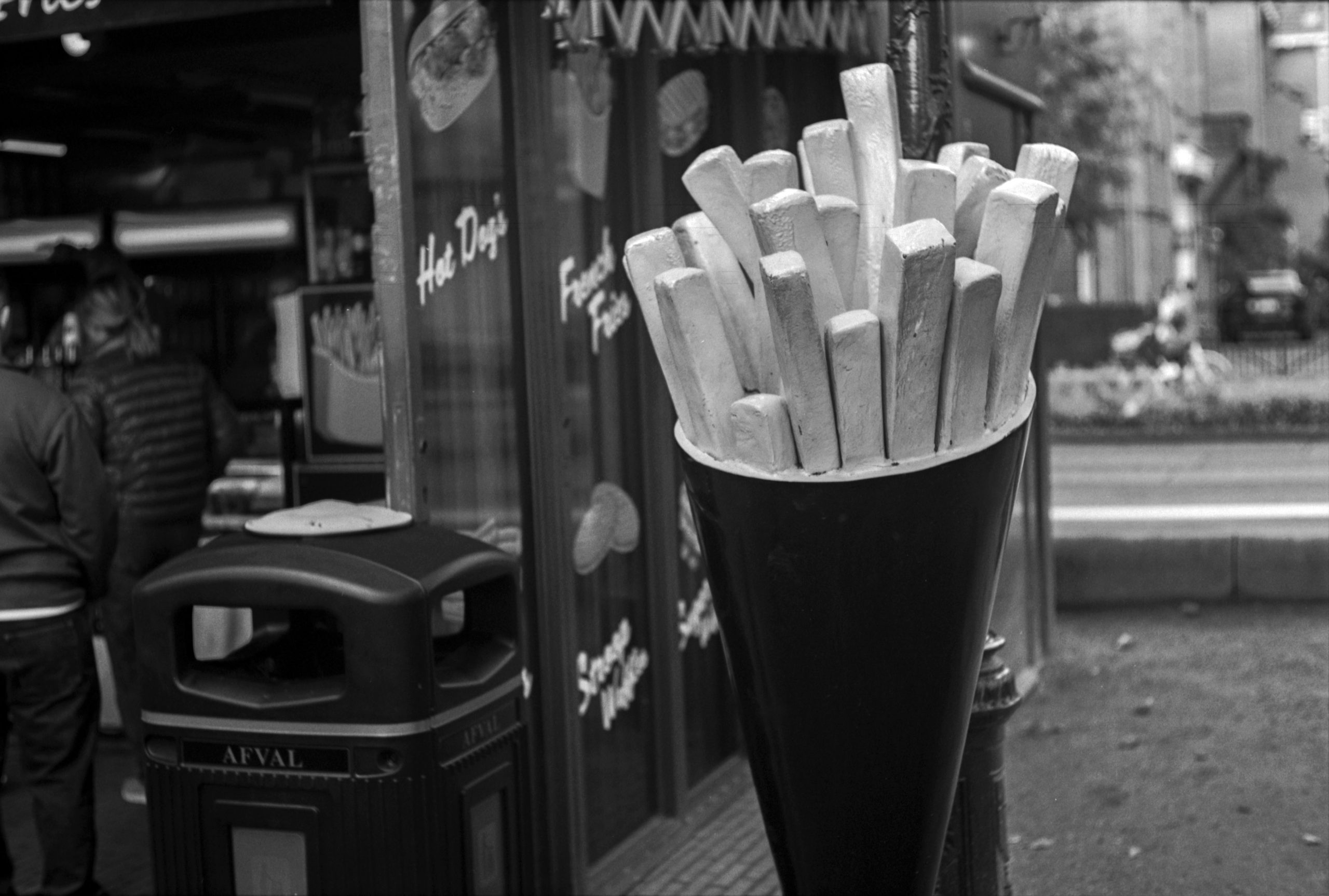 A close-up of a cone filled with unseasoned French fries, placed in front of a restaurant with people in the background.