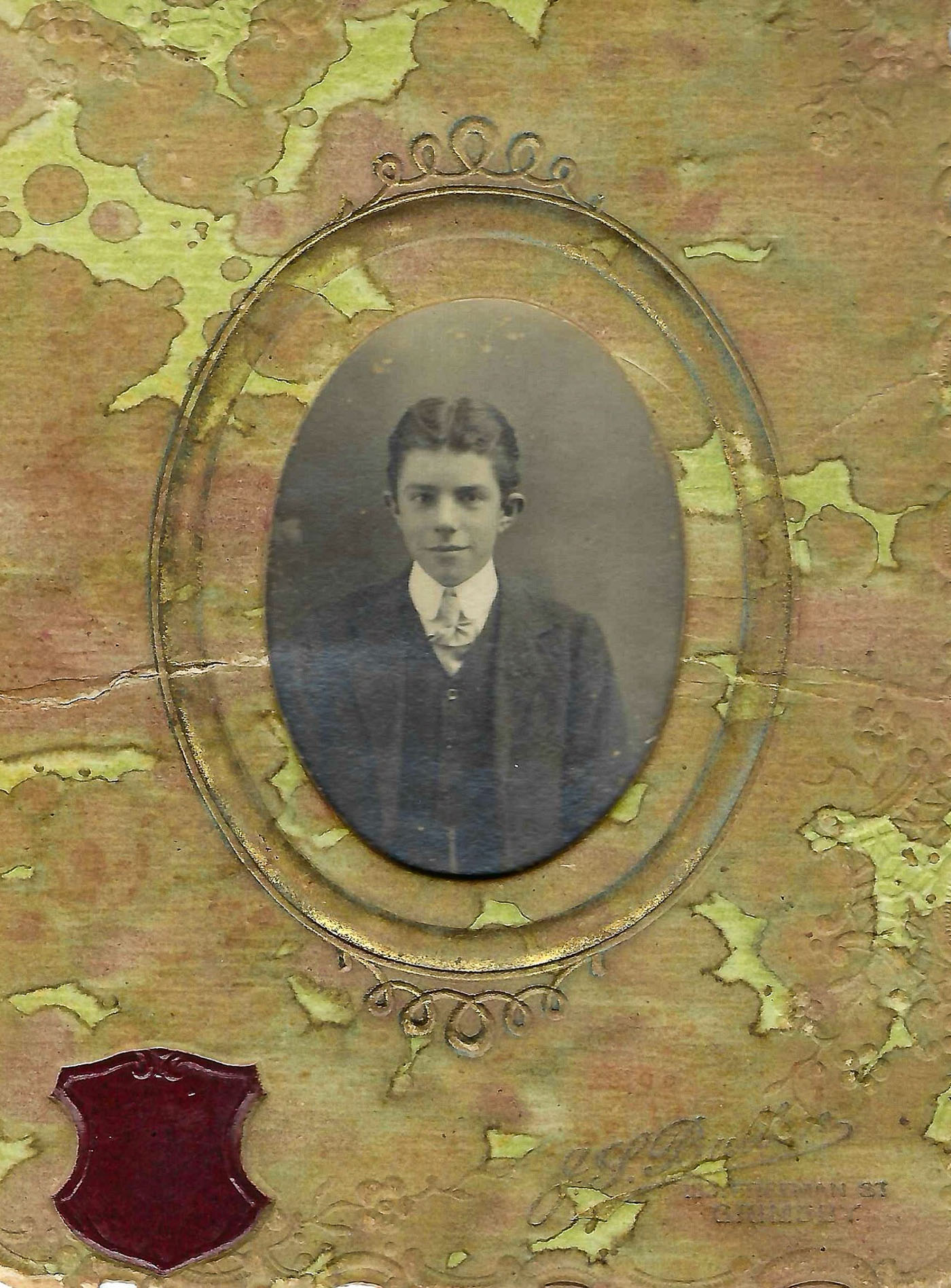 A vintage portrait of a young man in formal attire, framed within an ornate design. The backdrop features a textured, greenish surface with intricate patterns, adding an artistic touch to the photograph.