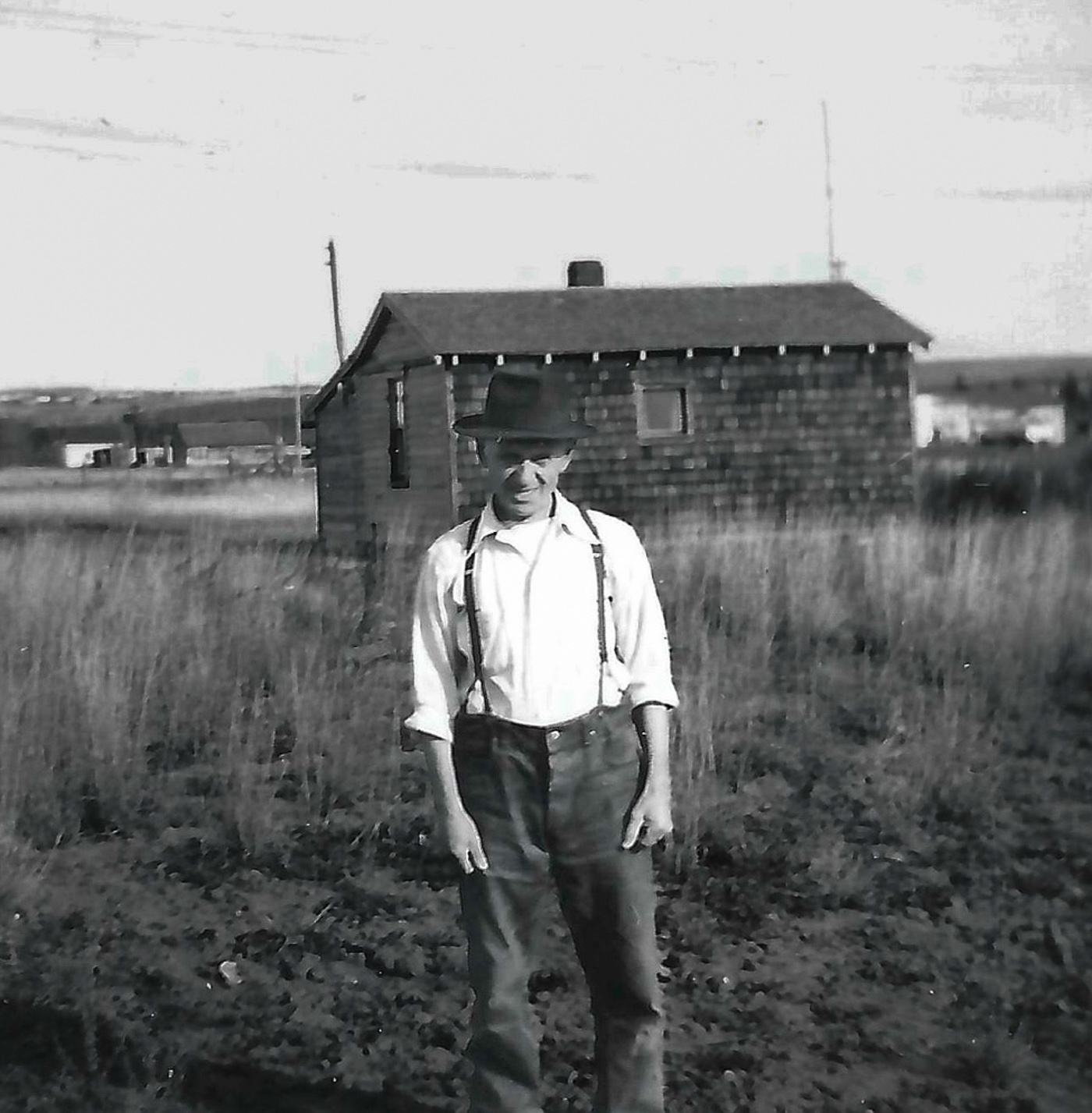 A historical black and white photograph of a man in suspenders and a hat, standing in a field with a wooden house in the background.