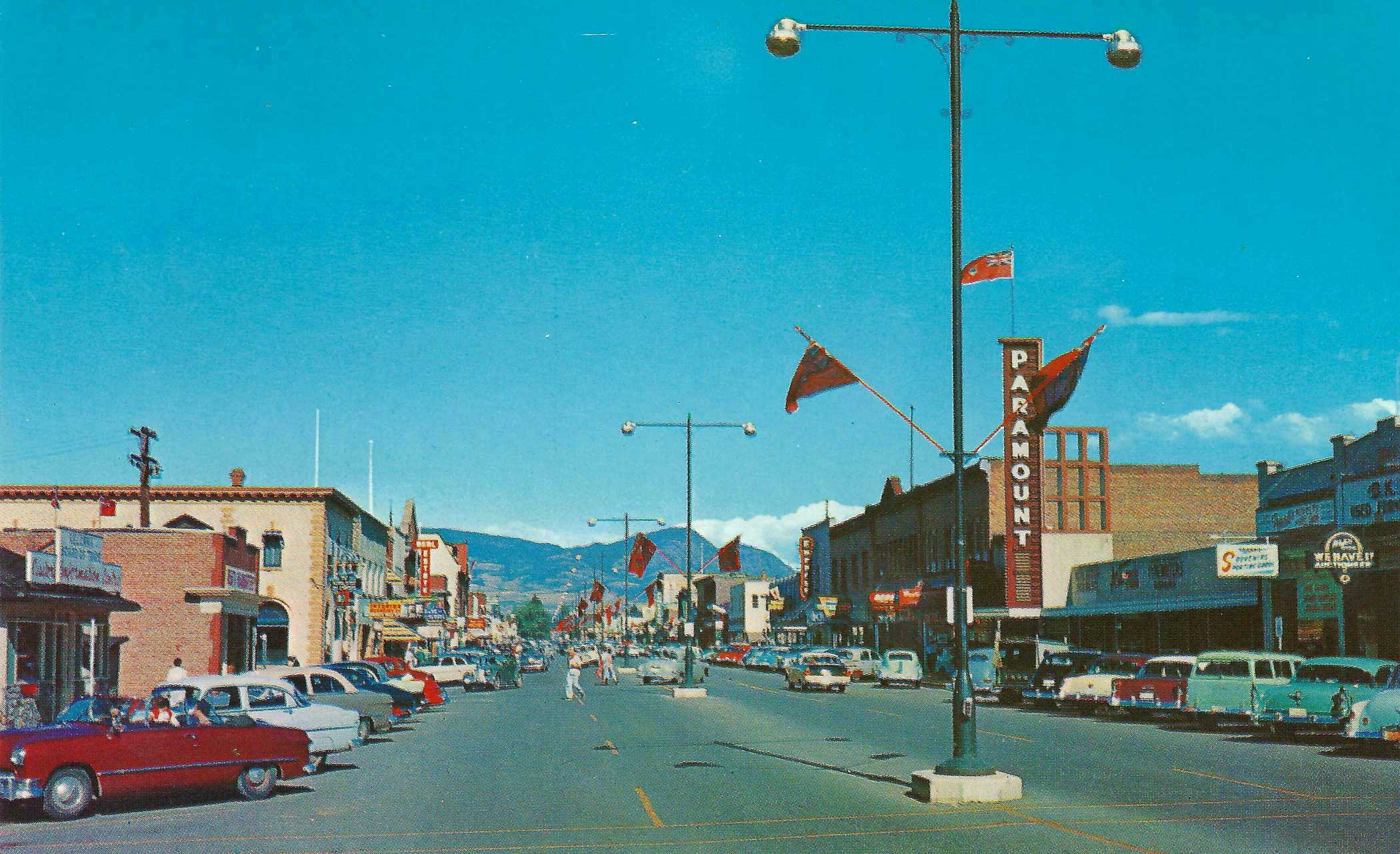 A vintage postcard-style image of a busy street in Kelowna, British Columbia, featuring parked cars and shops, with blue skies and mountains in the background.