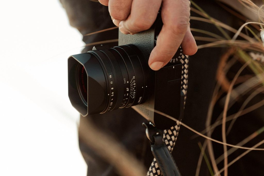 A close-up of a person holding the Leica Q3 Monochrom camera, featuring its lens and black body design against a blurred background.