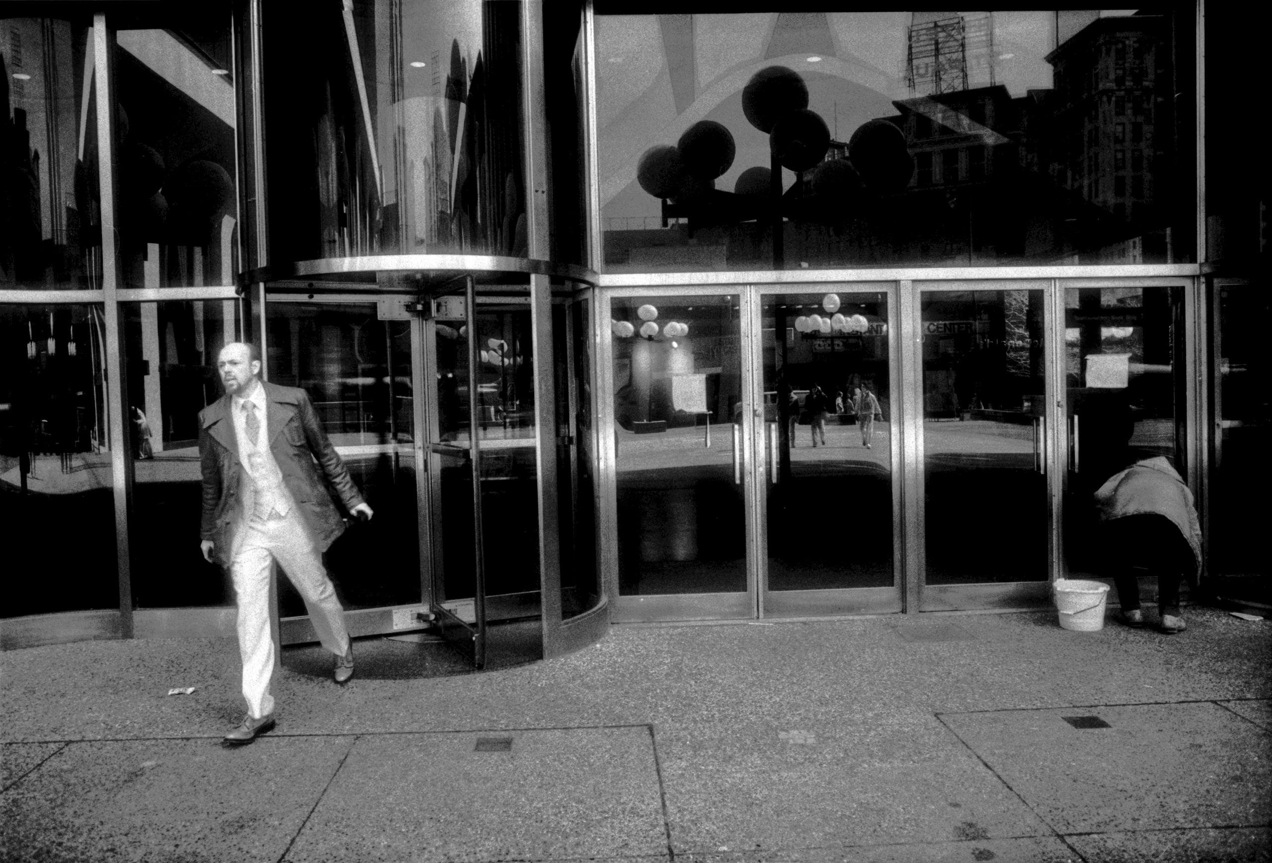 A man in a suit exits a building with mirrored glass doors, while another figure crouches near the entrance, possibly cleaning. The scene captures urban life in black and white.
