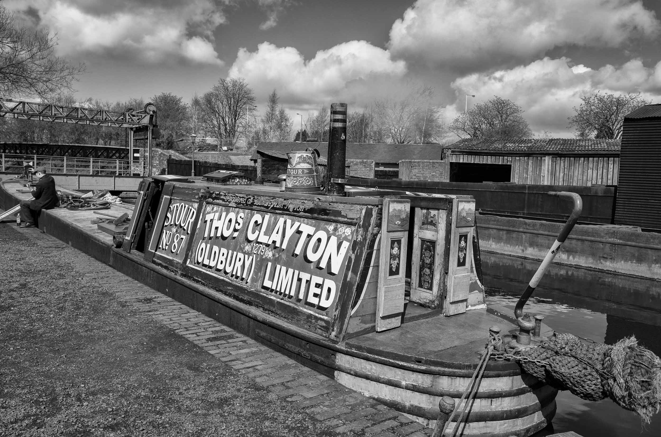 A black and white photograph of a barge named 'THOS CLAYTON OLD BURY LIMITED' docked at a canal, with trees and buildings in the background under a cloudy sky.