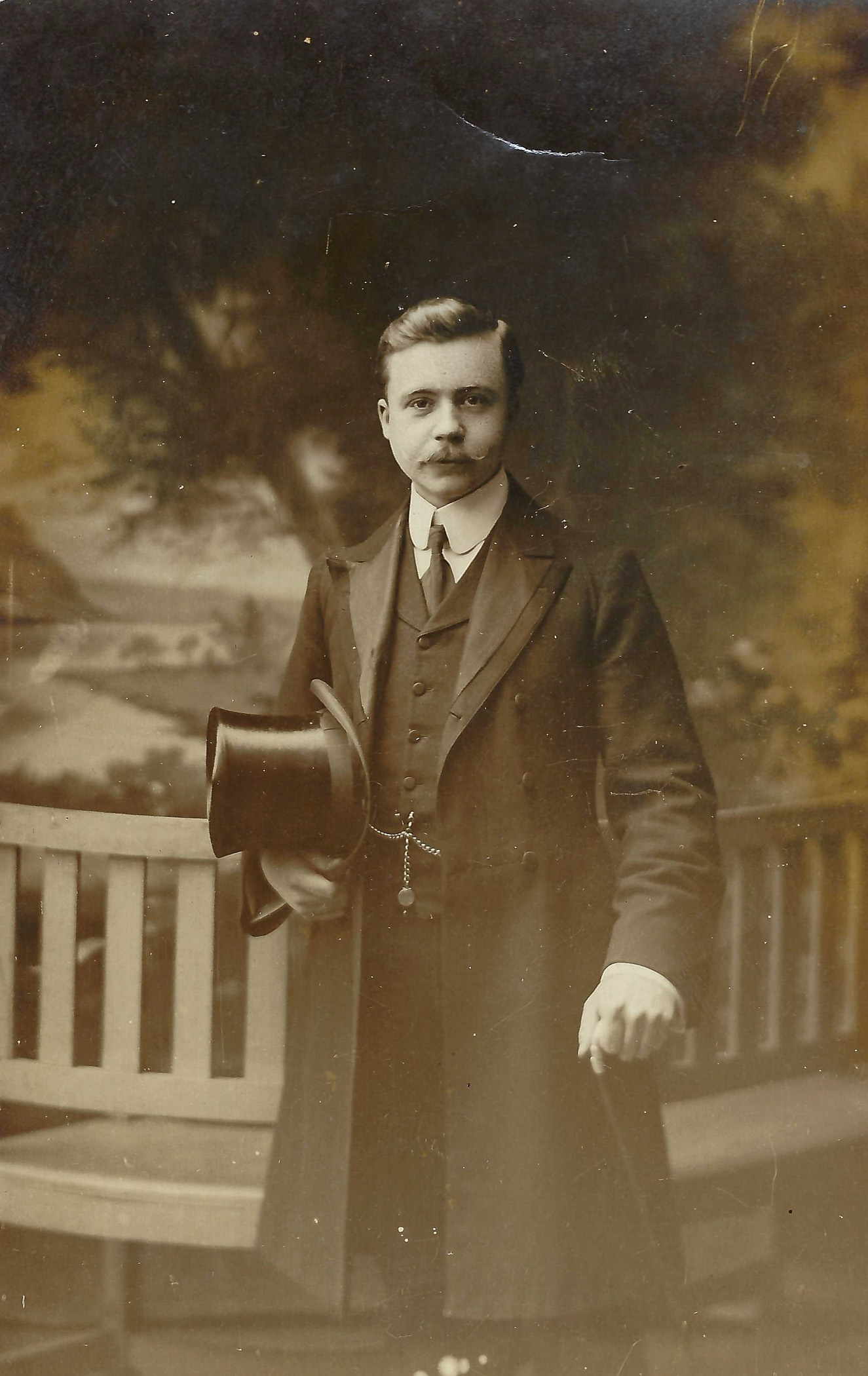 A vintage photograph of a young man dressed in formal attire, holding a top hat. He has a mustache and appears to be posing outdoors, possibly in a garden setting.