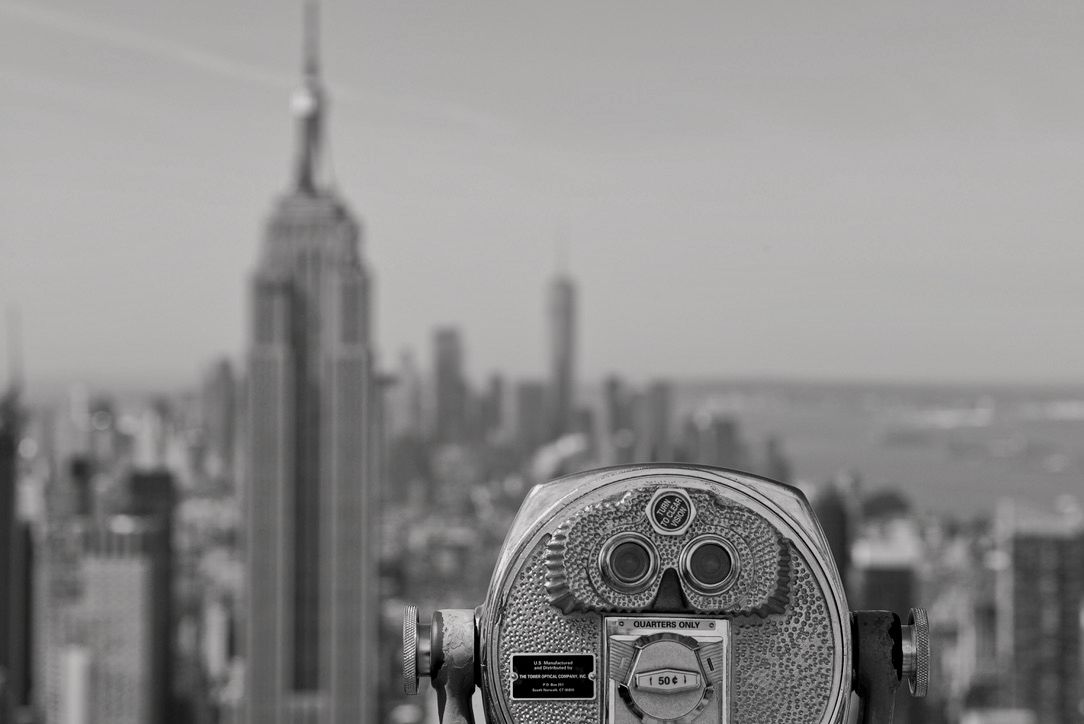 A black and white image of a binocular viewer overlooking a city skyline featuring the Empire State Building in the background.