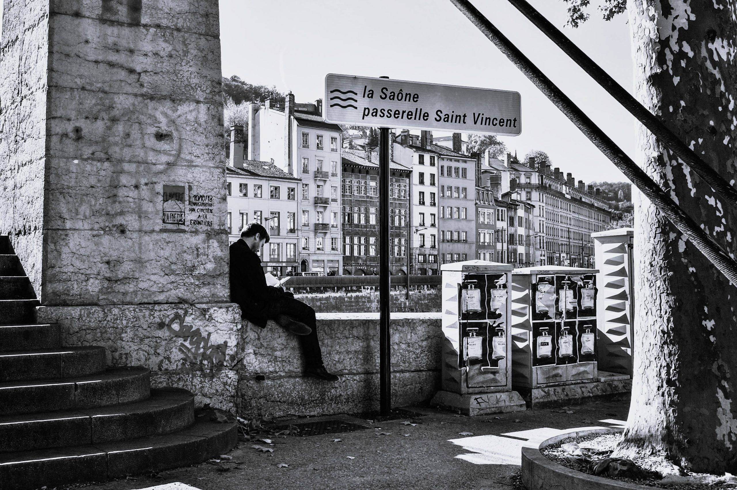 A person sitting on steps near the Saône River, looking at their phone, with historical buildings in the background and a sign indicating passerelle Saint Vincent.