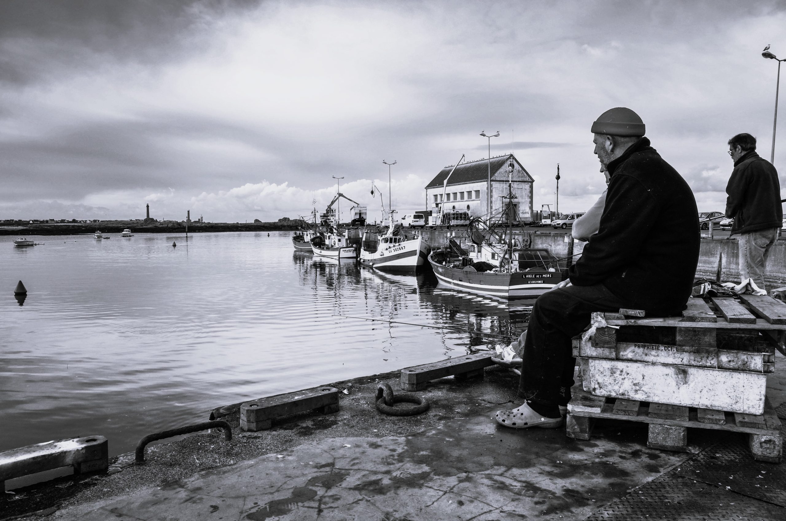 A black and white photograph depicting a man sitting on a dock, gazing at a calm harbor with fishing boats and a building in the background under a cloudy sky.