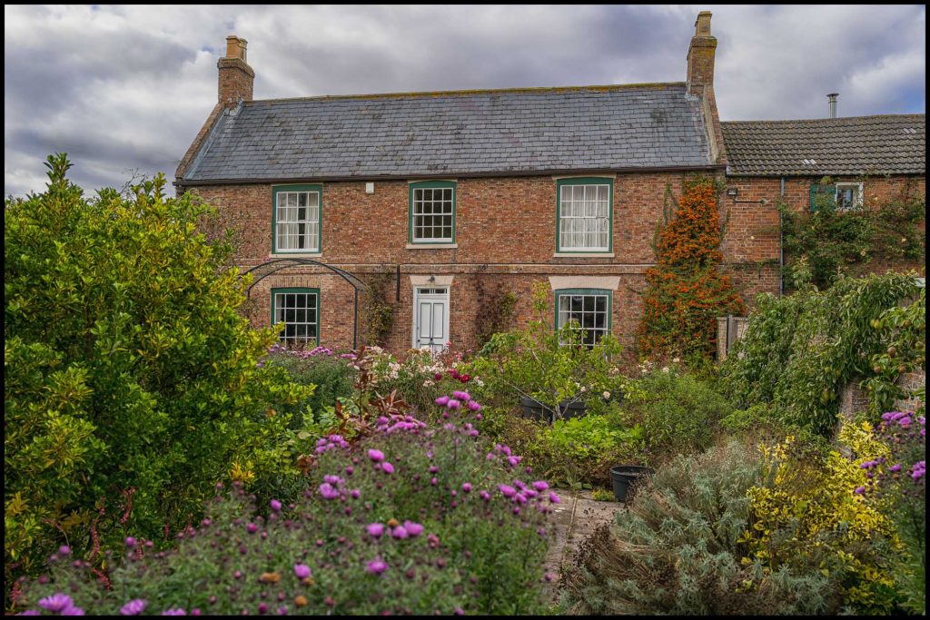 A charming brick house surrounded by a vibrant garden filled with colorful flowers and greenery under a cloudy sky.