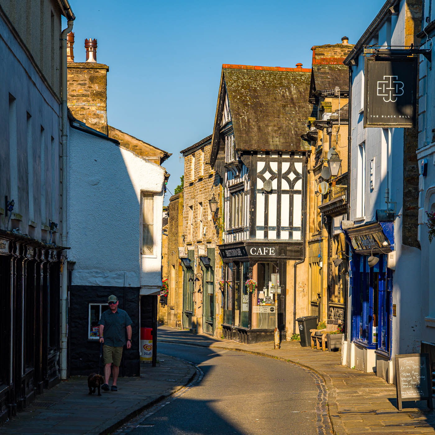 A picturesque street scene in a Yorkshire village, showcasing traditional stone buildings with colorful shop fronts, a cafe, and a man walking a dog under clear blue skies.