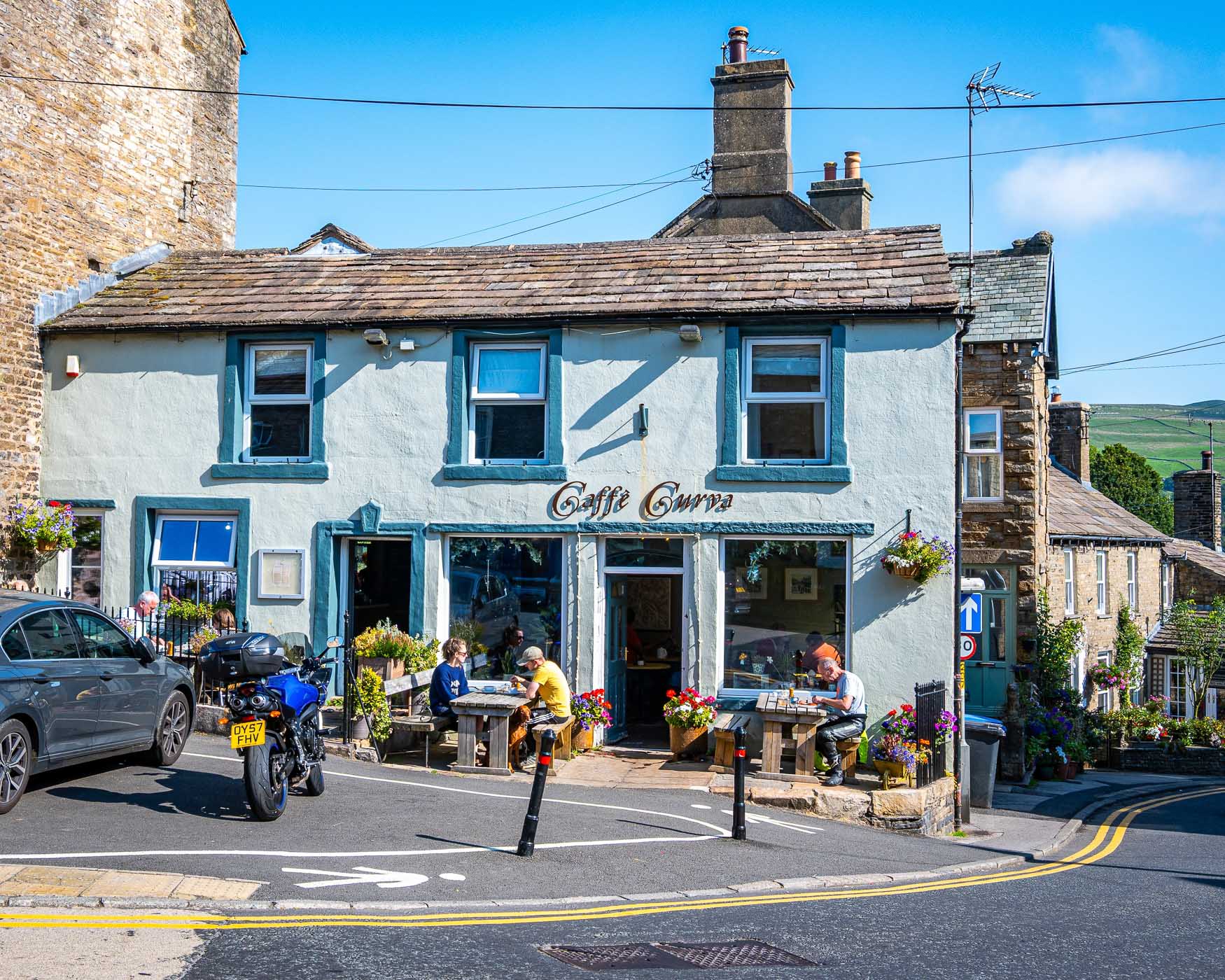 A charming café named 'Caffè Curra' located in a picturesque stone building, with outdoor seating adorned with flowers. Patrons are seen enjoying their meals, while a motorcycle and a car are parked nearby against a clear blue sky.