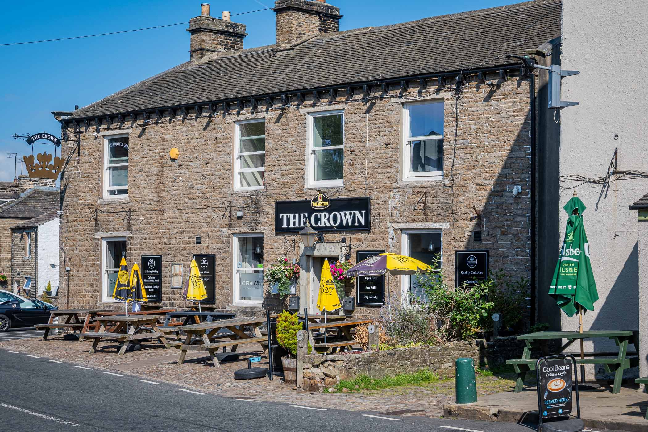 Exterior view of The Crown pub, a traditional stone building with patio seating and umbrellas, located on a sunny street in Yorkshire.