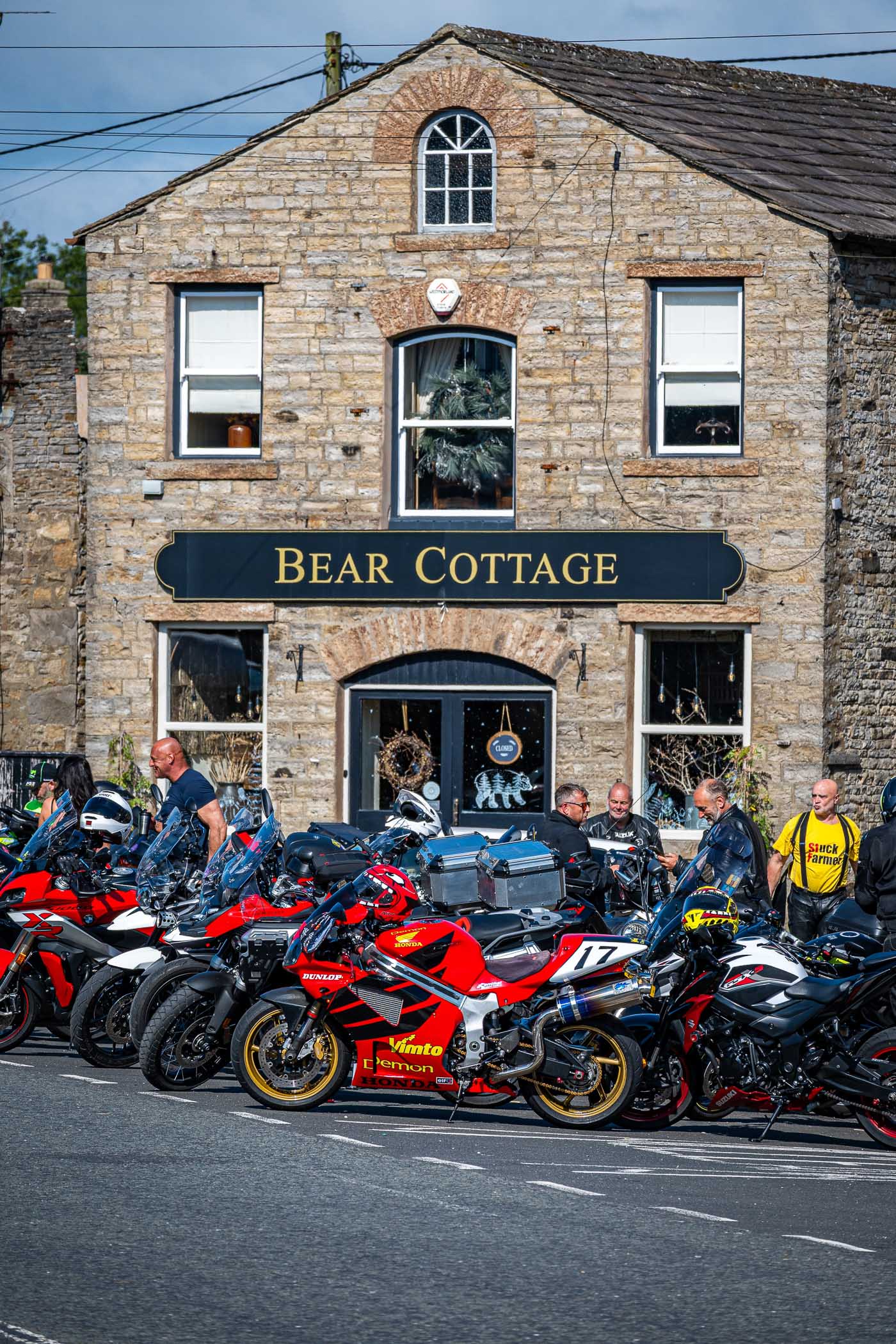 A group of motorcyclists gathered outside Bear Cottage, an old stone building, with several colorful motorcycles parked in the foreground. The scene captures a sunny day, highlighting the lively atmosphere among riders.