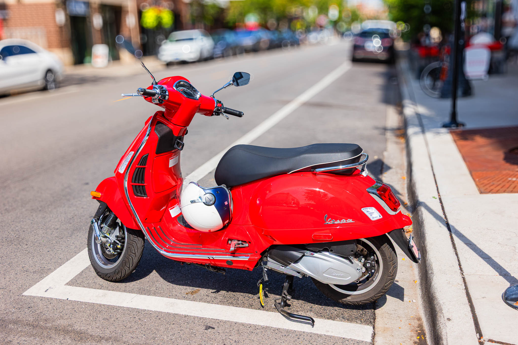 A red Vespa scooter parked on a city street, showcasing its sleek design against a blurred backdrop of parked cars and greenery.