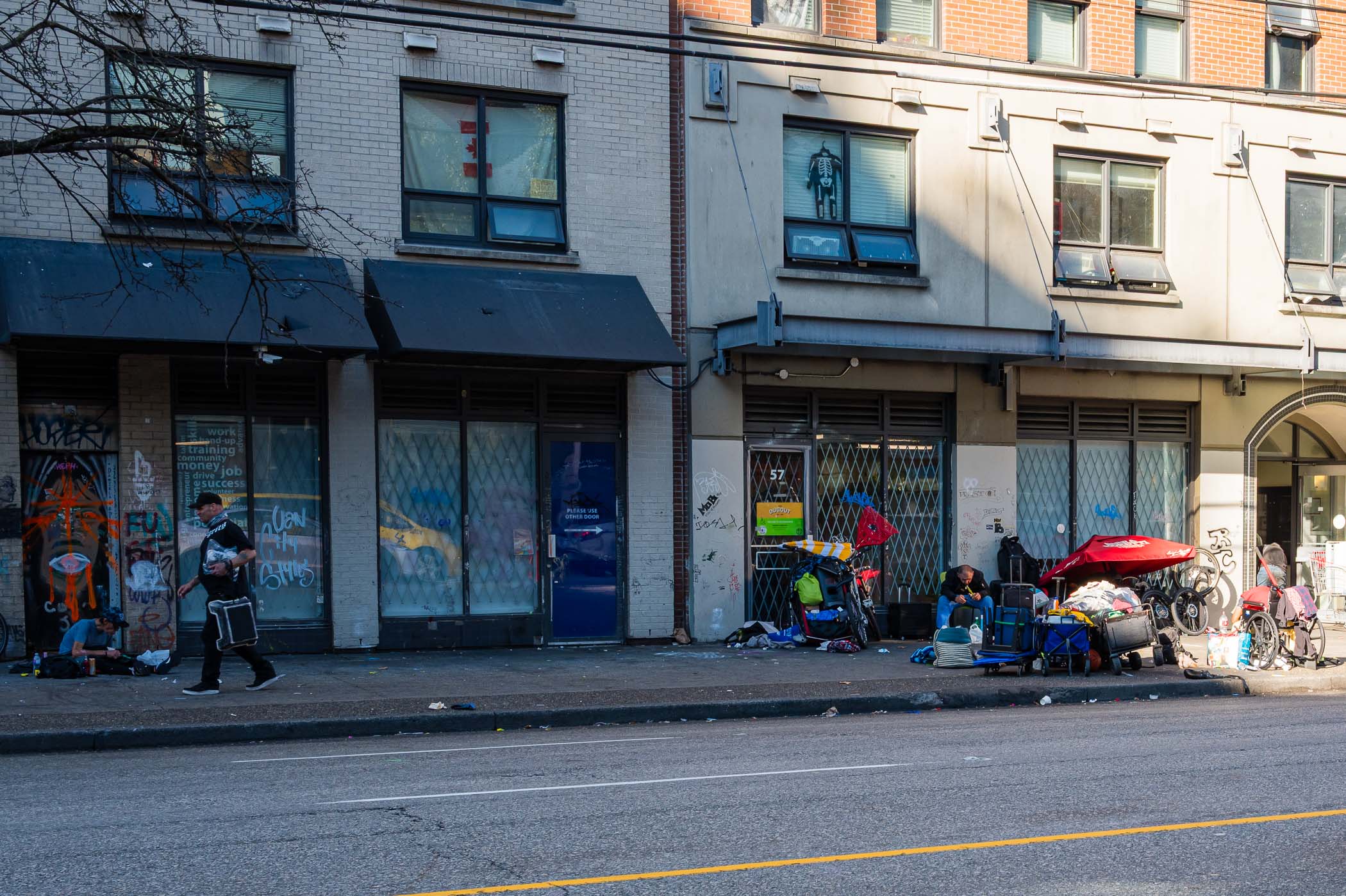 Street scene in Vancouver showing a homeless encampment with individuals, bicycles, and belongings beside a building with graffiti.