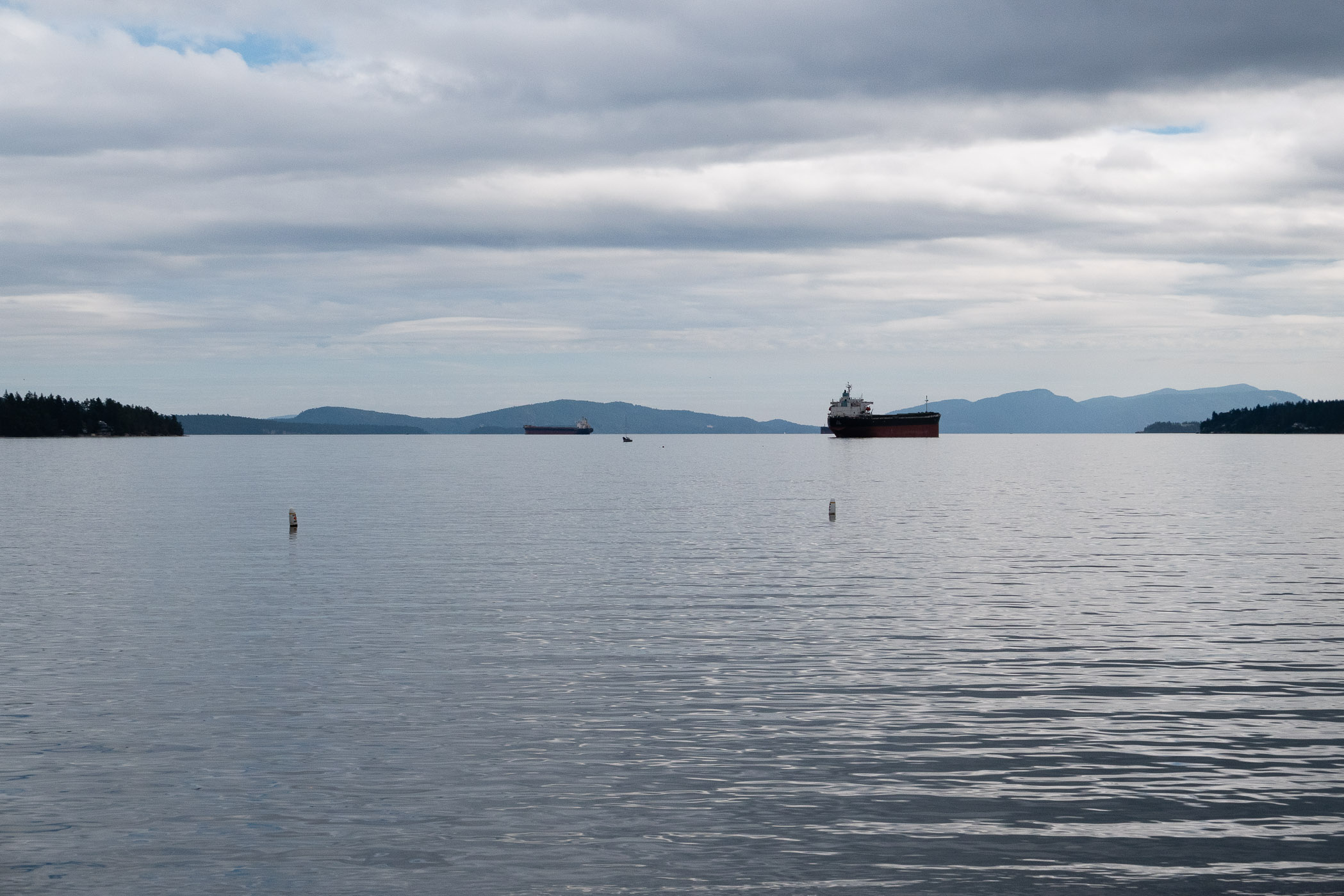 A tranquil view of a harbor with two ships anchored on calm waters, surrounded by distant mountains and a cloudy sky.