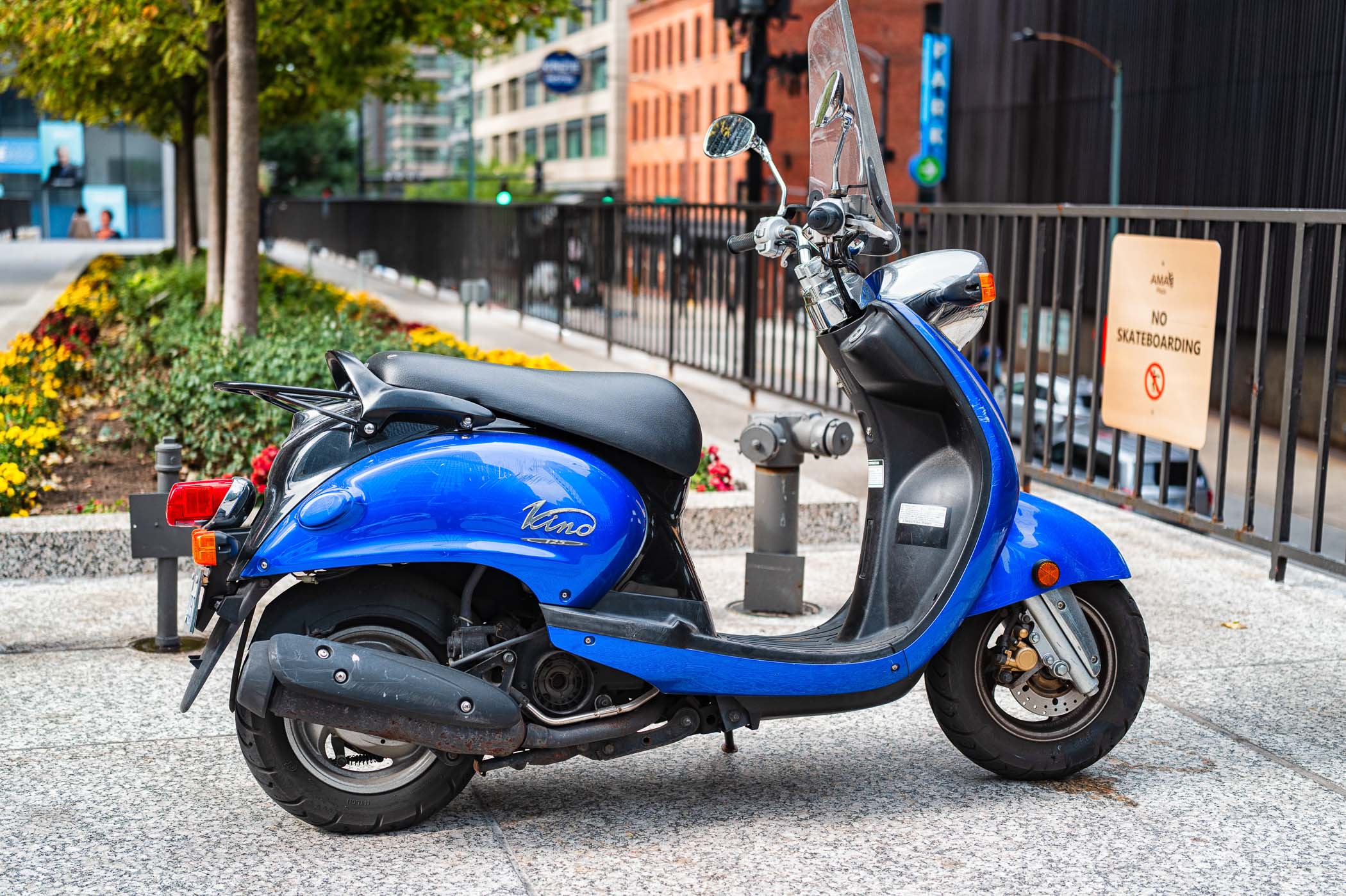 A blue Vespa scooter parked on a city street, surrounded by colorful flowers and urban scenery.