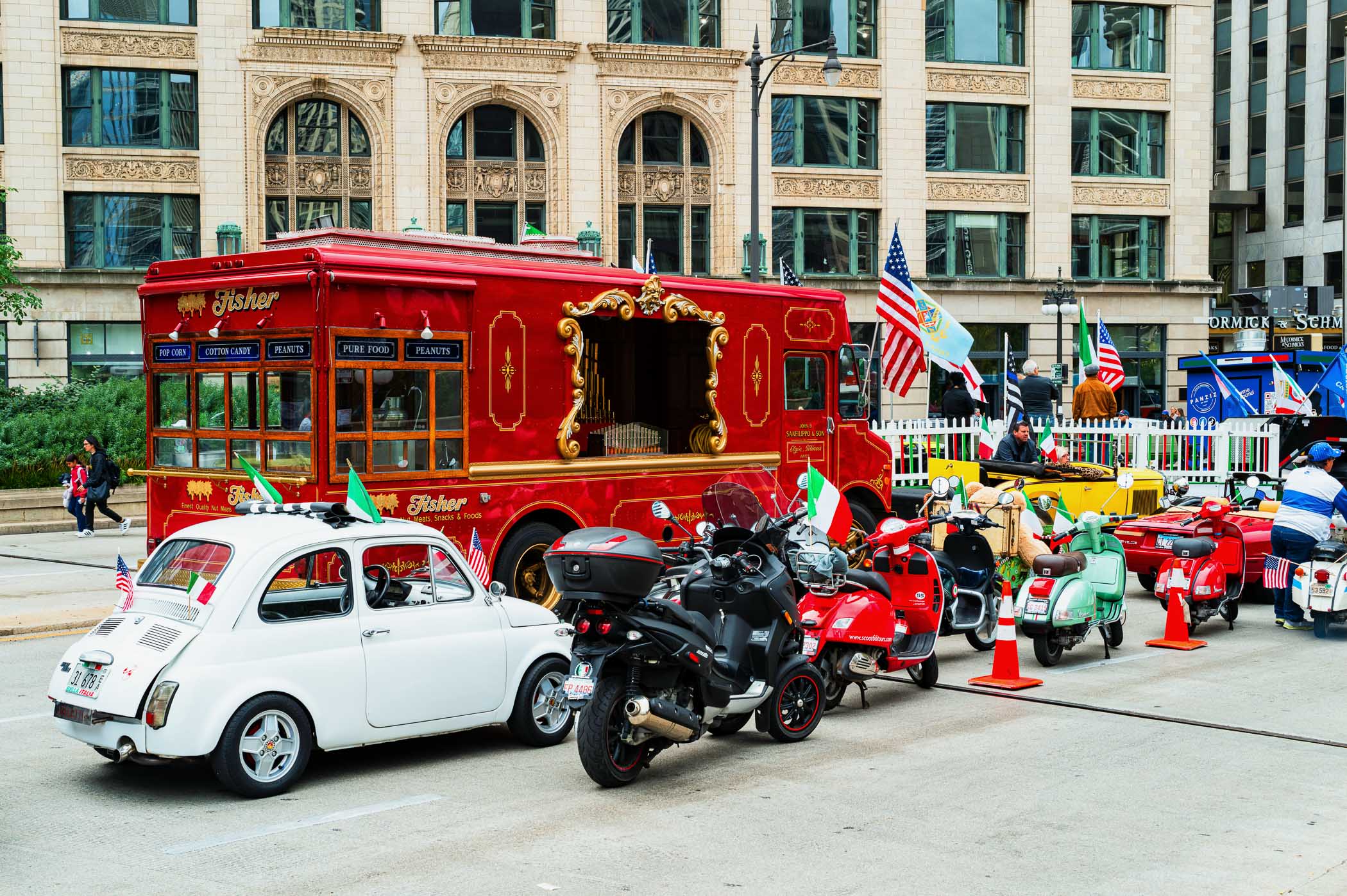 A vibrant scene from Columbus Day in Chicago featuring a red food truck decorated with gold accents, alongside a collection of vintage scooters and an iconic white Fiat car, all adorned with flags, capturing the festive atmosphere.