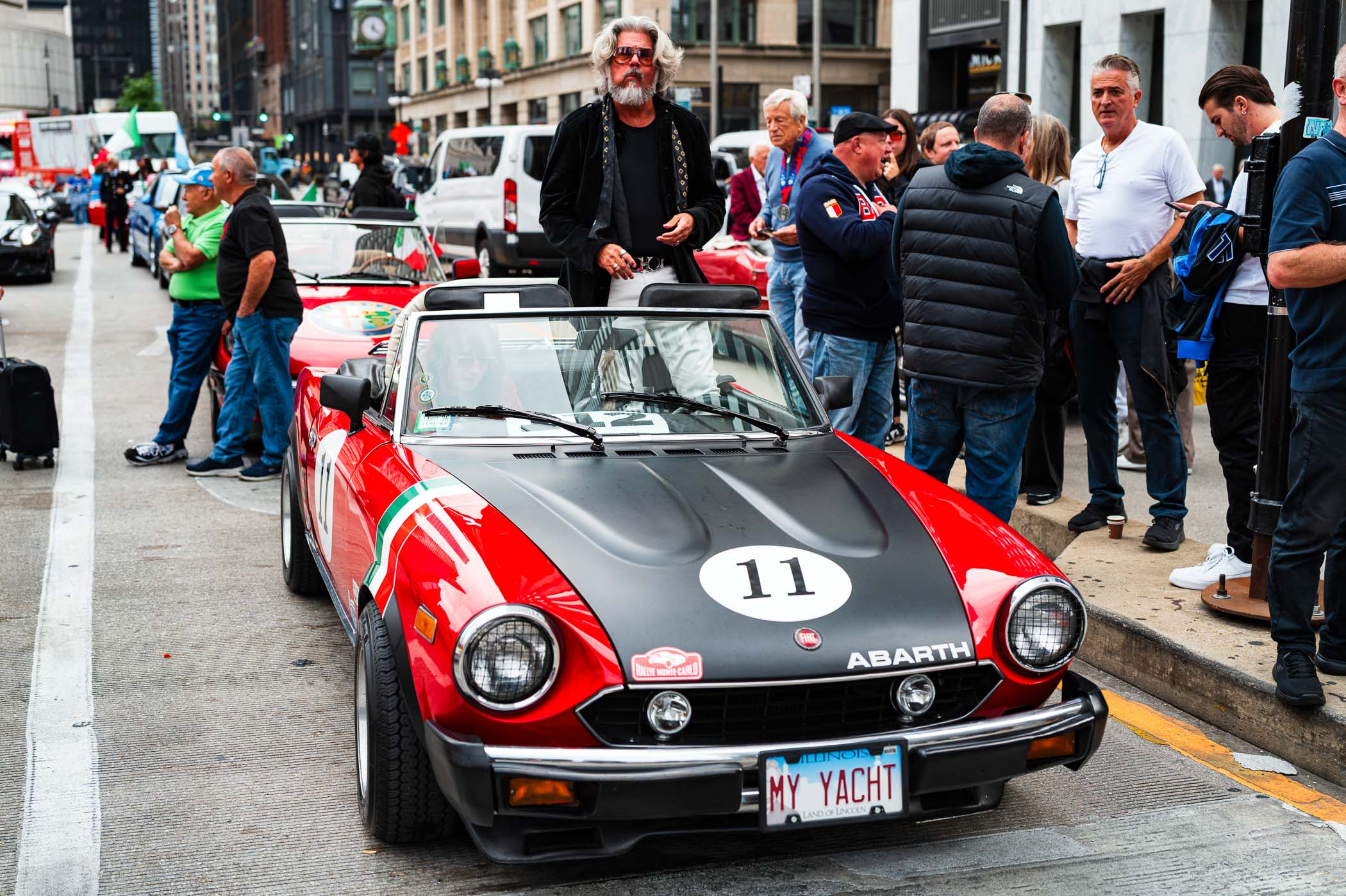 A red Abarth car with a black hood and the number 11, parked along a street during a Columbus Day celebration in Chicago, surrounded by a crowd of people in casual attire.