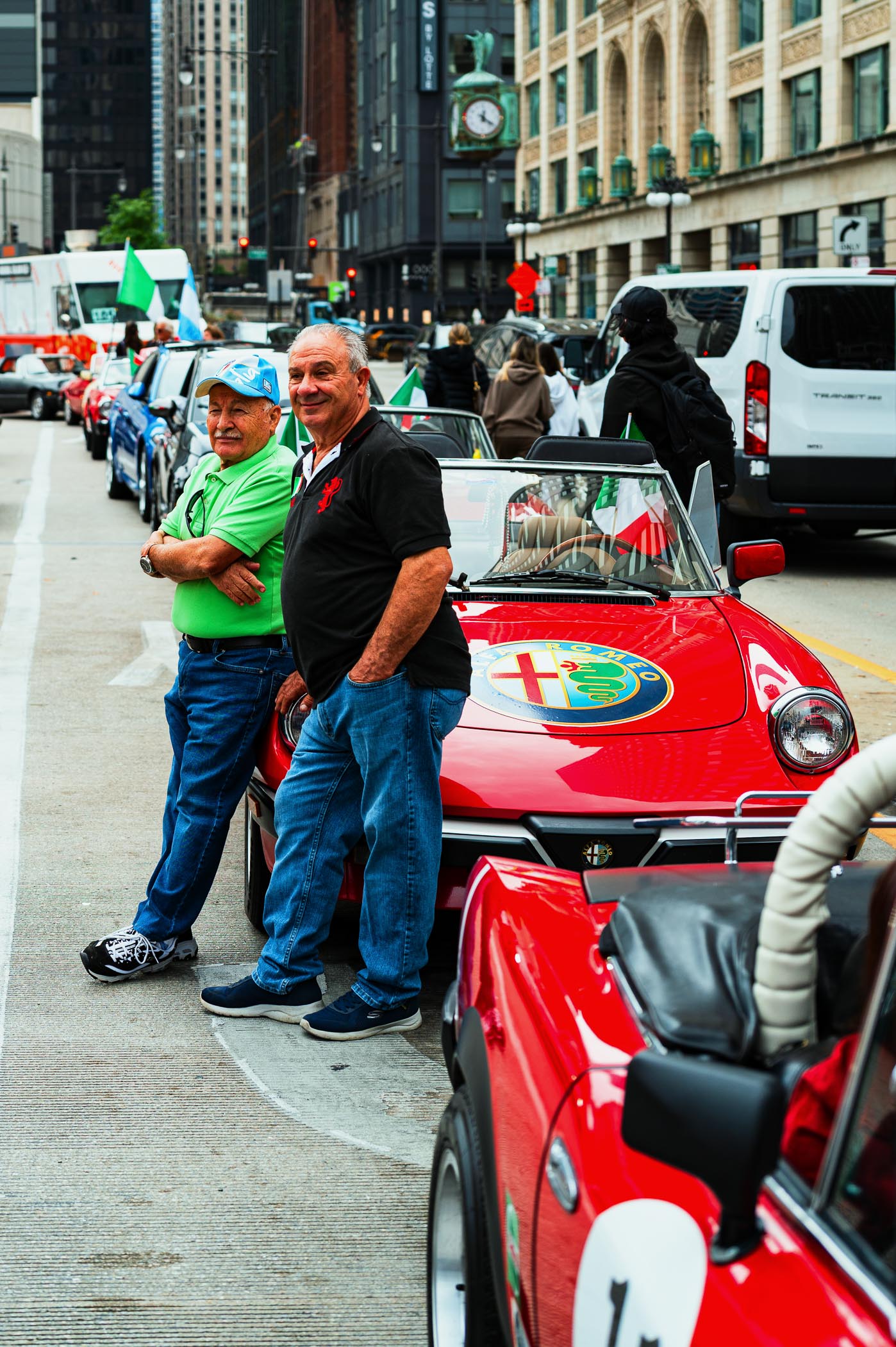 Two men pose near a red Alfa Romeo convertible decorated with Italian flags during the Columbus Day parade in Chicago, surrounded by a lineup of cars and urban buildings.