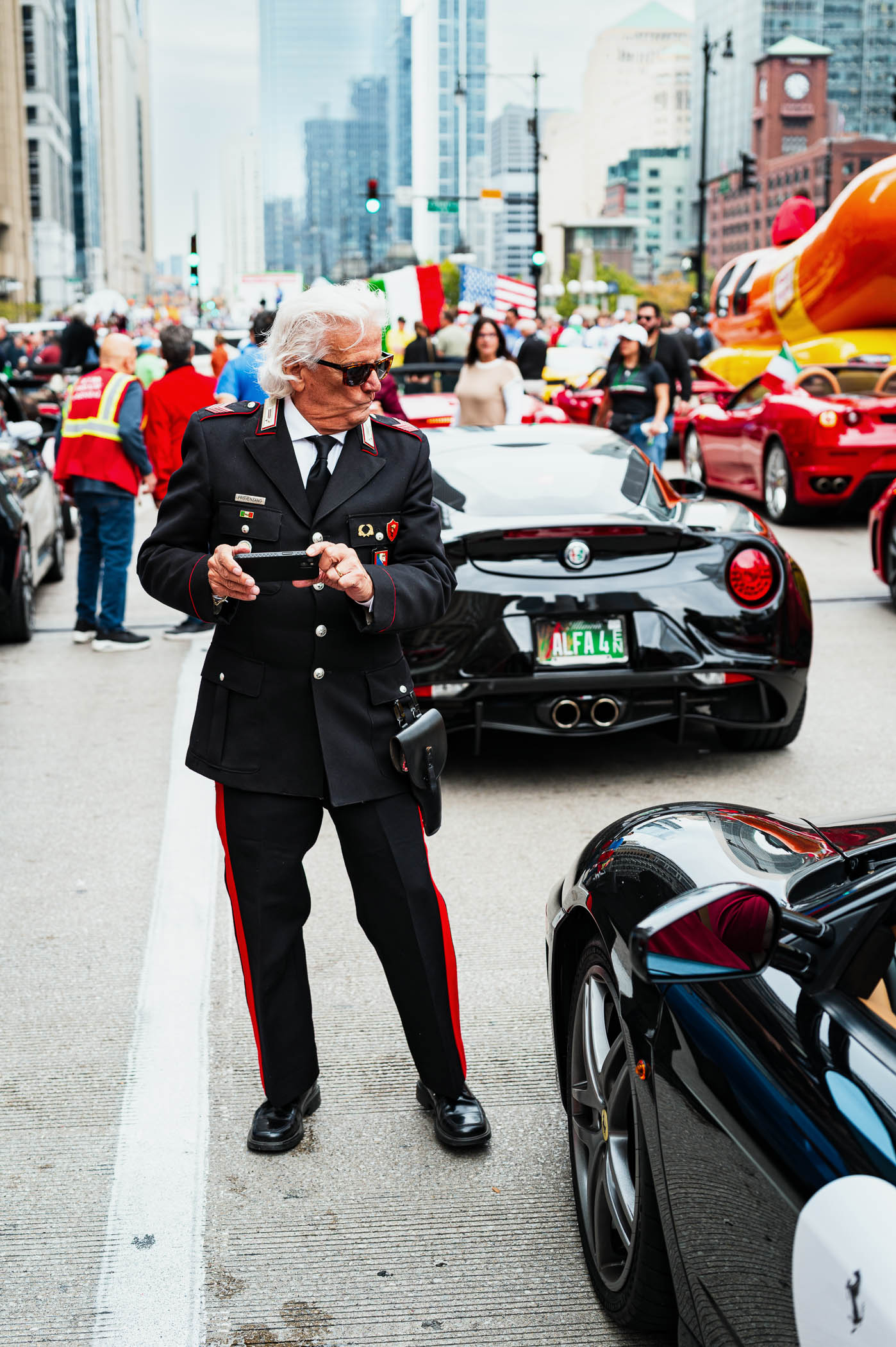 An older man in a black police uniform with red accents stands on a busy street during a festive event in Chicago. He holds a device and appears to be observing the surroundings, while colorful cars, people, and flags decorate the scene.