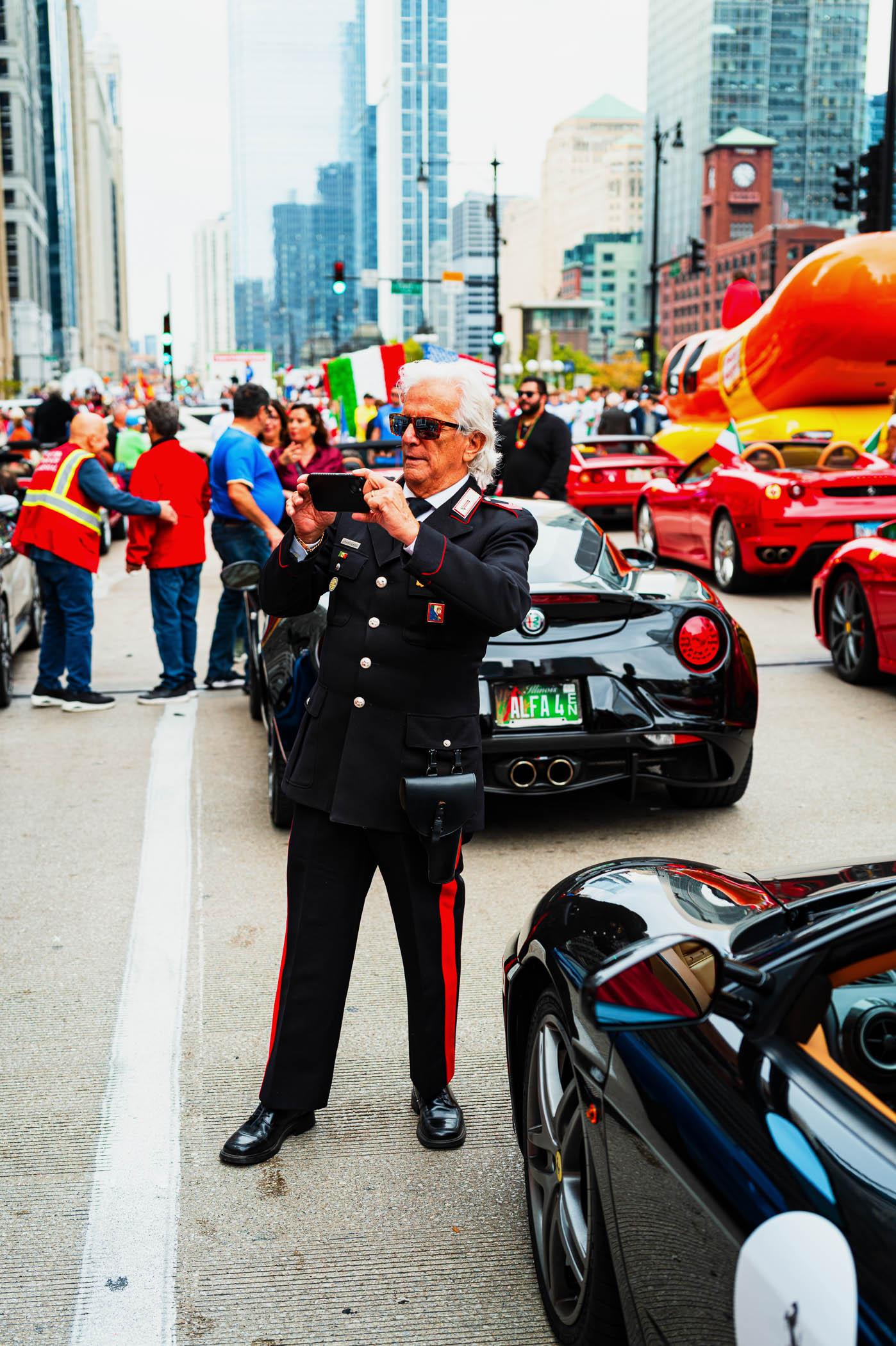 An older man dressed in a black police uniform with red accents stands in a street filled with people and classic cars. He is holding a smartphone and taking a photo, while colorful Italian flags adorn the scene.